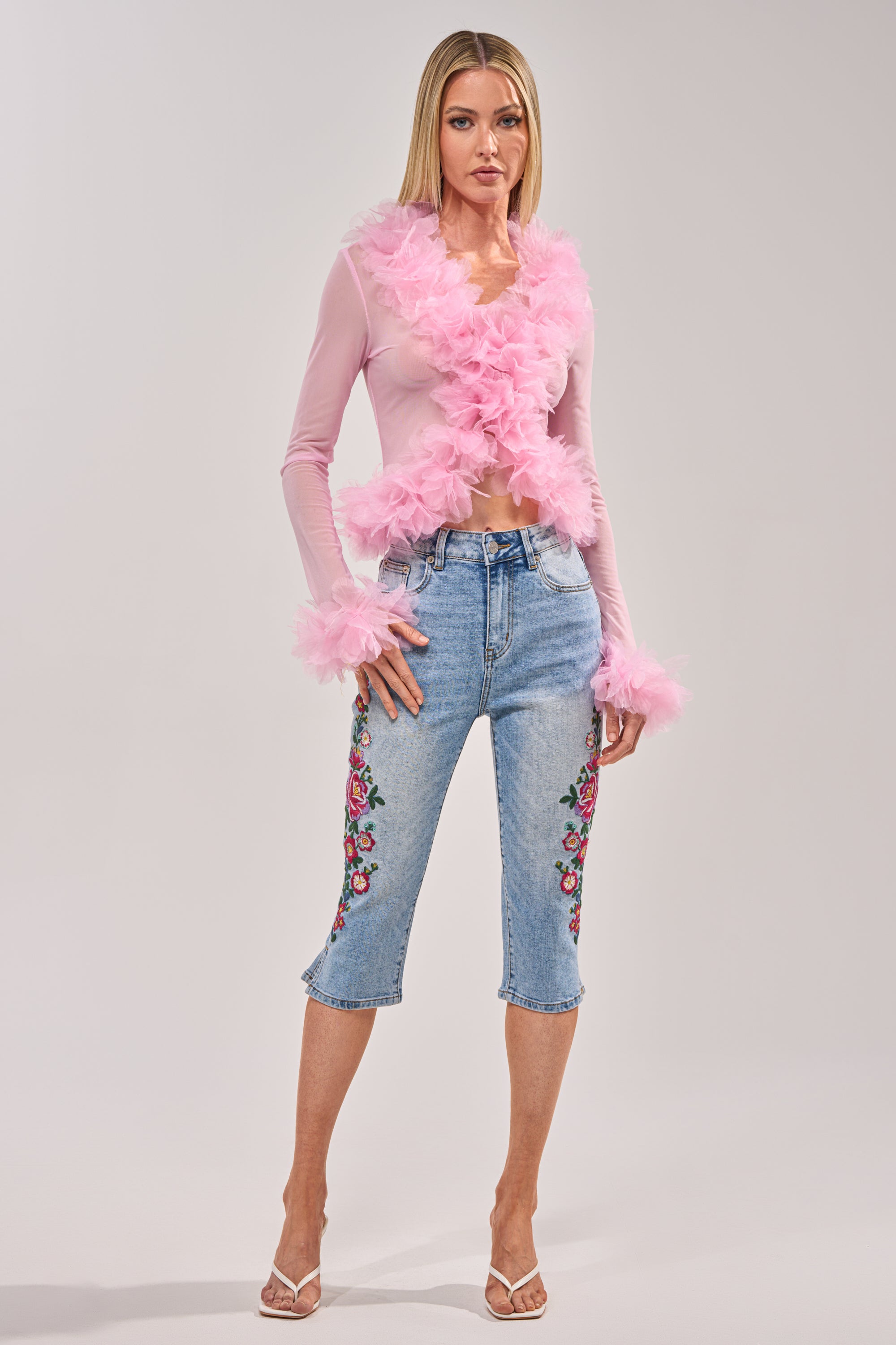 A woman wearing the GIVE ME FLOWERS FLORAL CAPRI, a sheer pink feather-trimmed top, and clear heeled sandals poses against a plain light background. 4