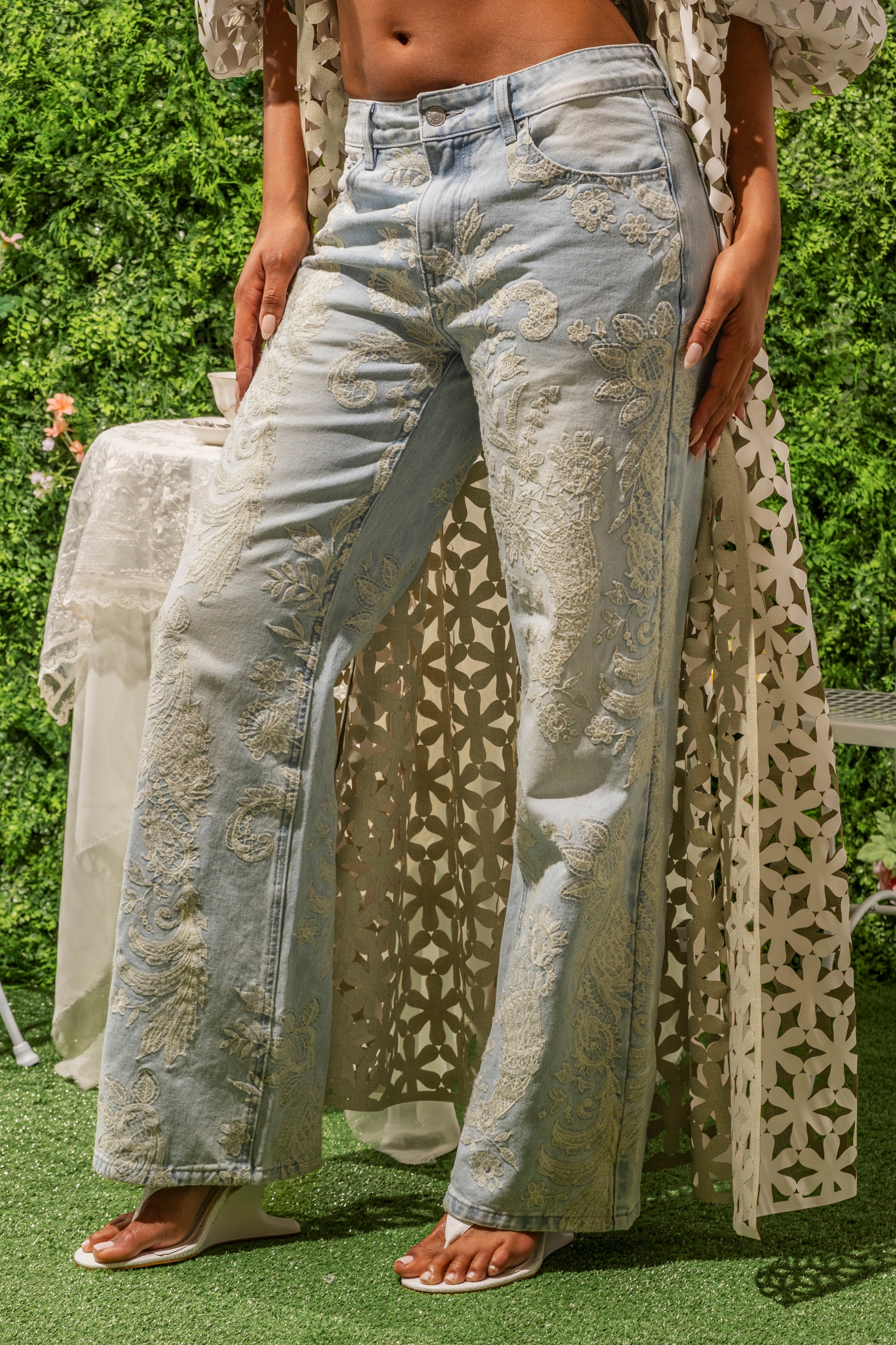 Wearing RODEO BABY DENIM light blue embroidered wide-leg jeans with floral accents and white sandals, a person stands outdoors on grass in a white lace cover-up, surrounded by greenery.