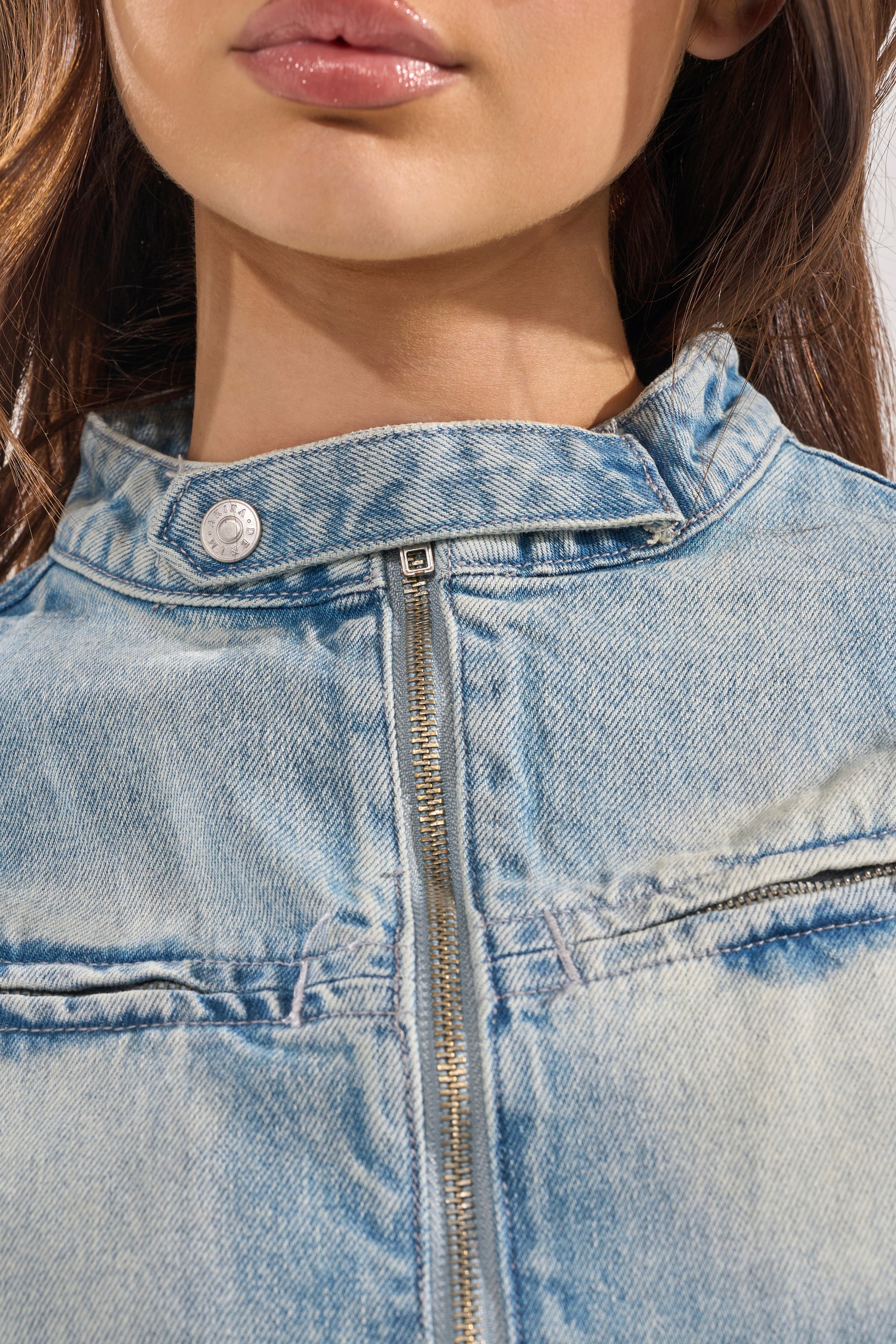 Close-up of a woman wearing the AFTER HOURS DENIM MOTO IN LIGHT BLUE, a cropped jacket featuring a front zipper and metal snap button at the collar. Only her chin, lips, and part of her hair are visible. 8