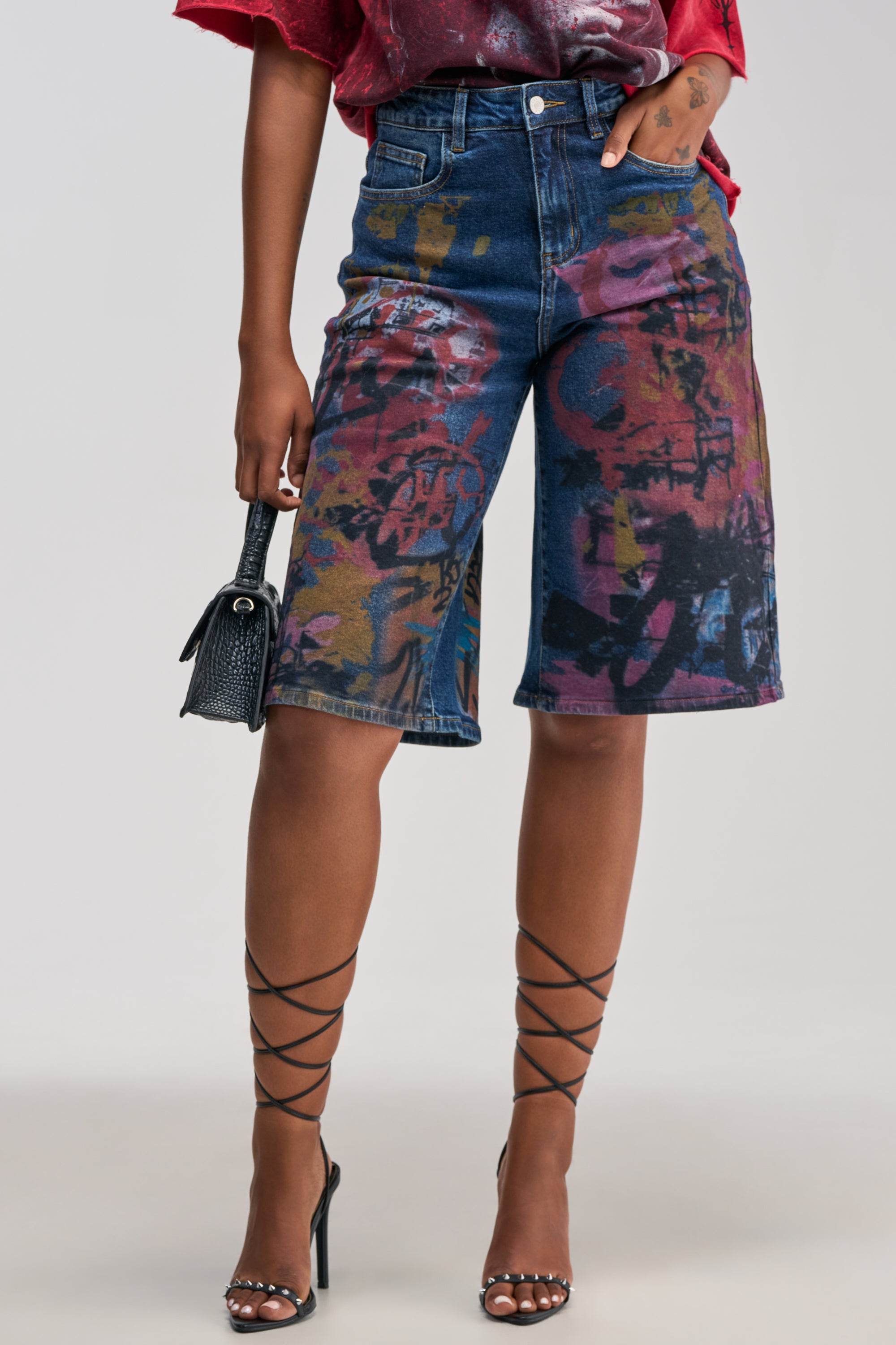Wearing the TRYING SOMETHING DIFF PAINTED DENIM Bermuda shorts, a person pairs the colorful, graffiti print denim with a red shirt, strappy high-heeled sandals, and a small black purse for a bold streetwear look. Only the lower body is visible.