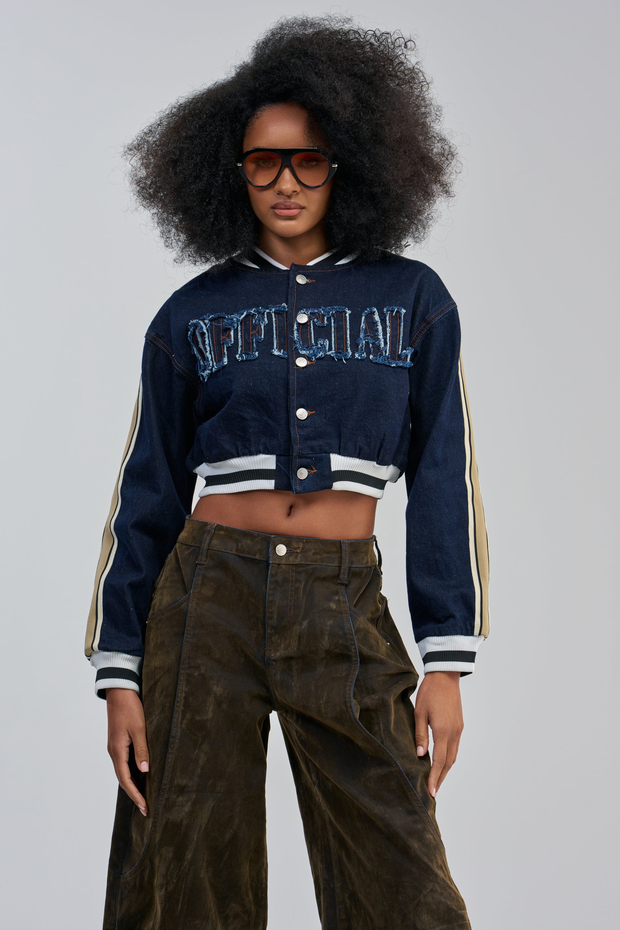 A woman with voluminous curly hair wears sunglasses, the OFFICIAL DENIM BOMBER—a navy cropped streetwear jacket featuring “OFFICIAL” text—and brown wide-leg pants, standing confidently against a plain background.
