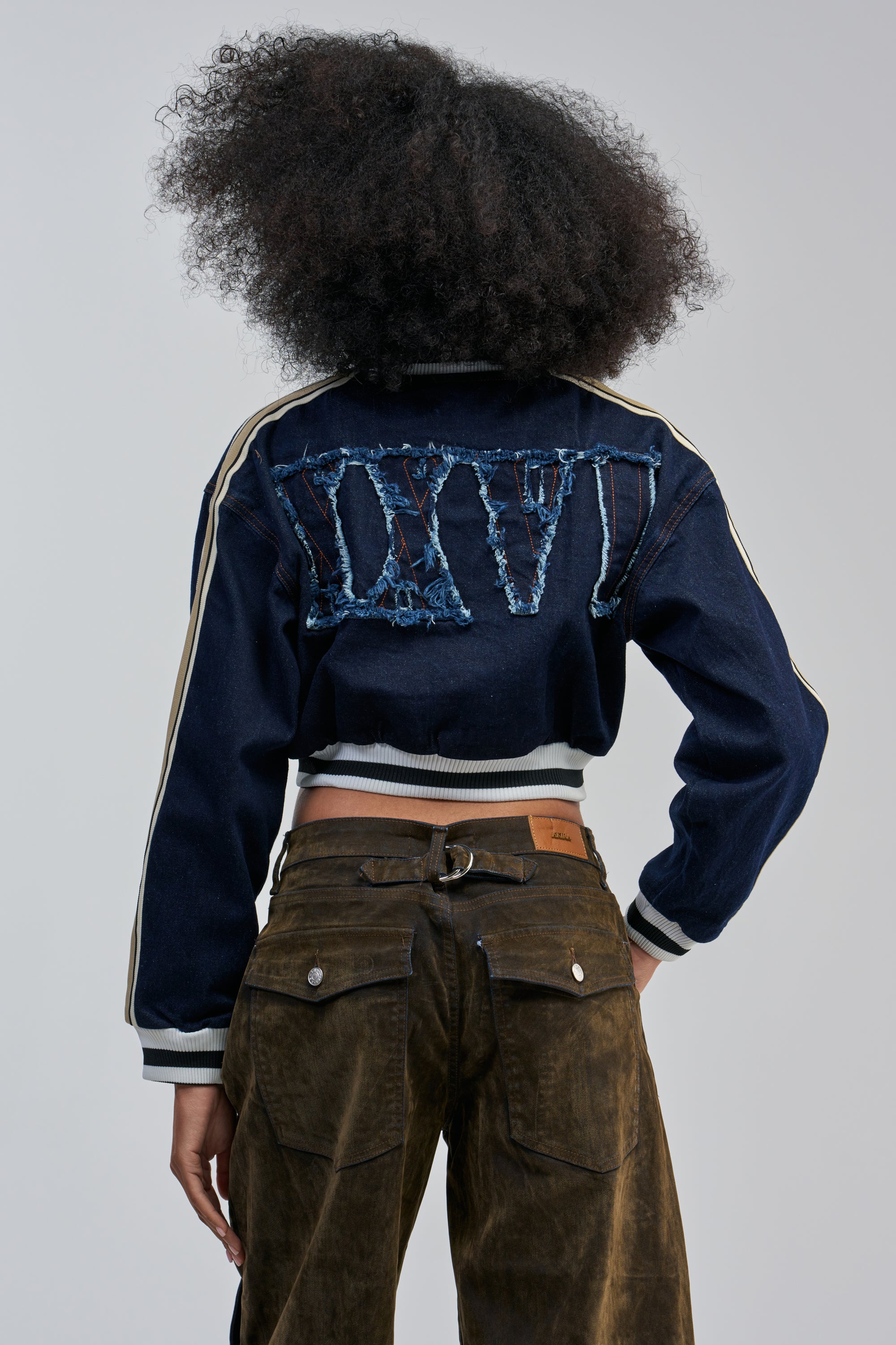 A person stands facing away, wearing the OFFICIAL DENIM BOMBER—a distressed denim streetwear jacket featuring bold Roman numerals “XXVI” on the back—paired with brown pants. Their voluminous, curly hair contrasts against a plain, light-colored background.