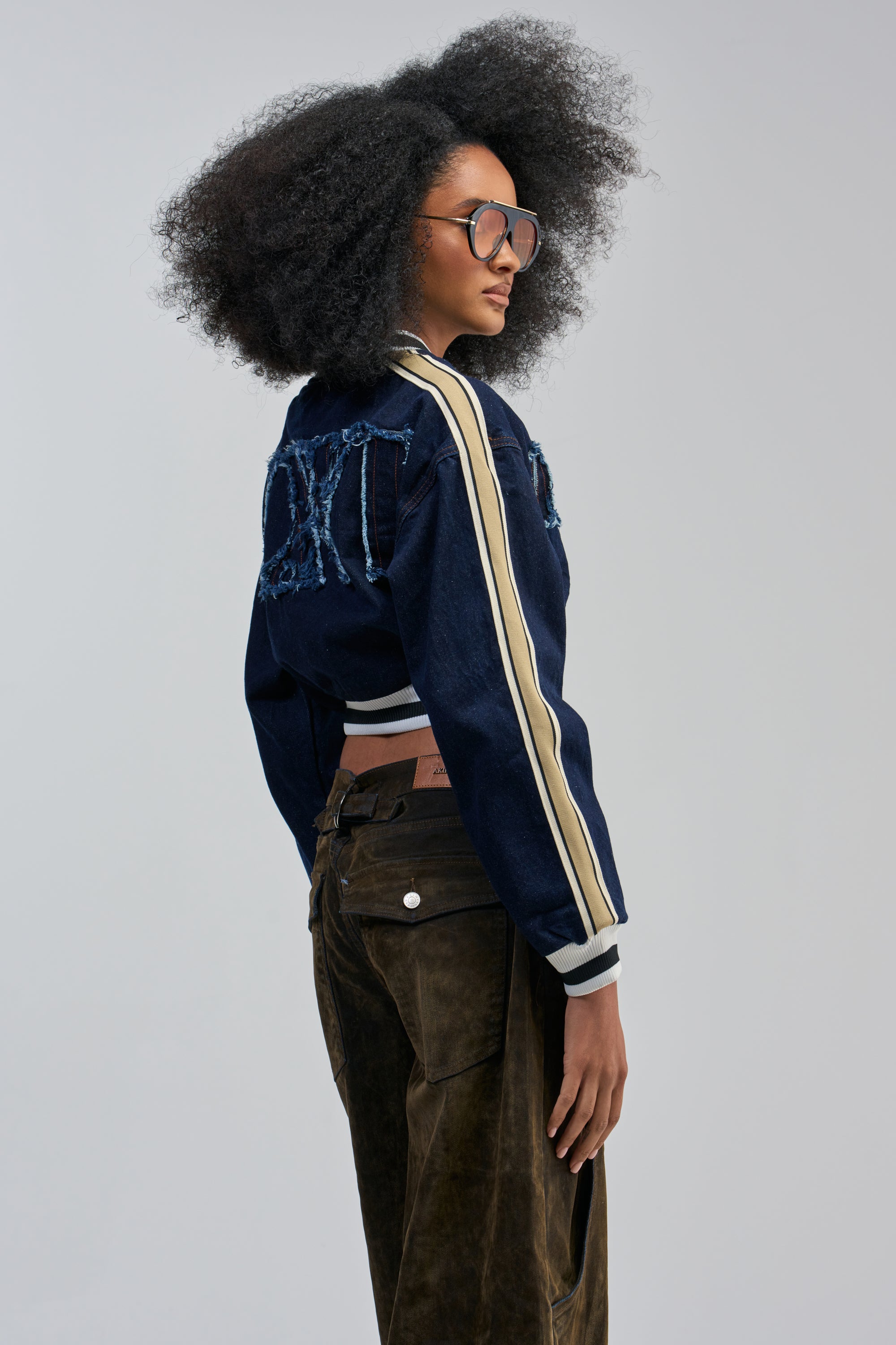 A woman with large, curly hair and oversized sunglasses wears the OFFICIAL DENIM BOMBER—a cropped dark wash denim jacket featuring frayed lettering on the back and striped sleeves—paired with dark brown suede pants, set against a plain light gray studio background.