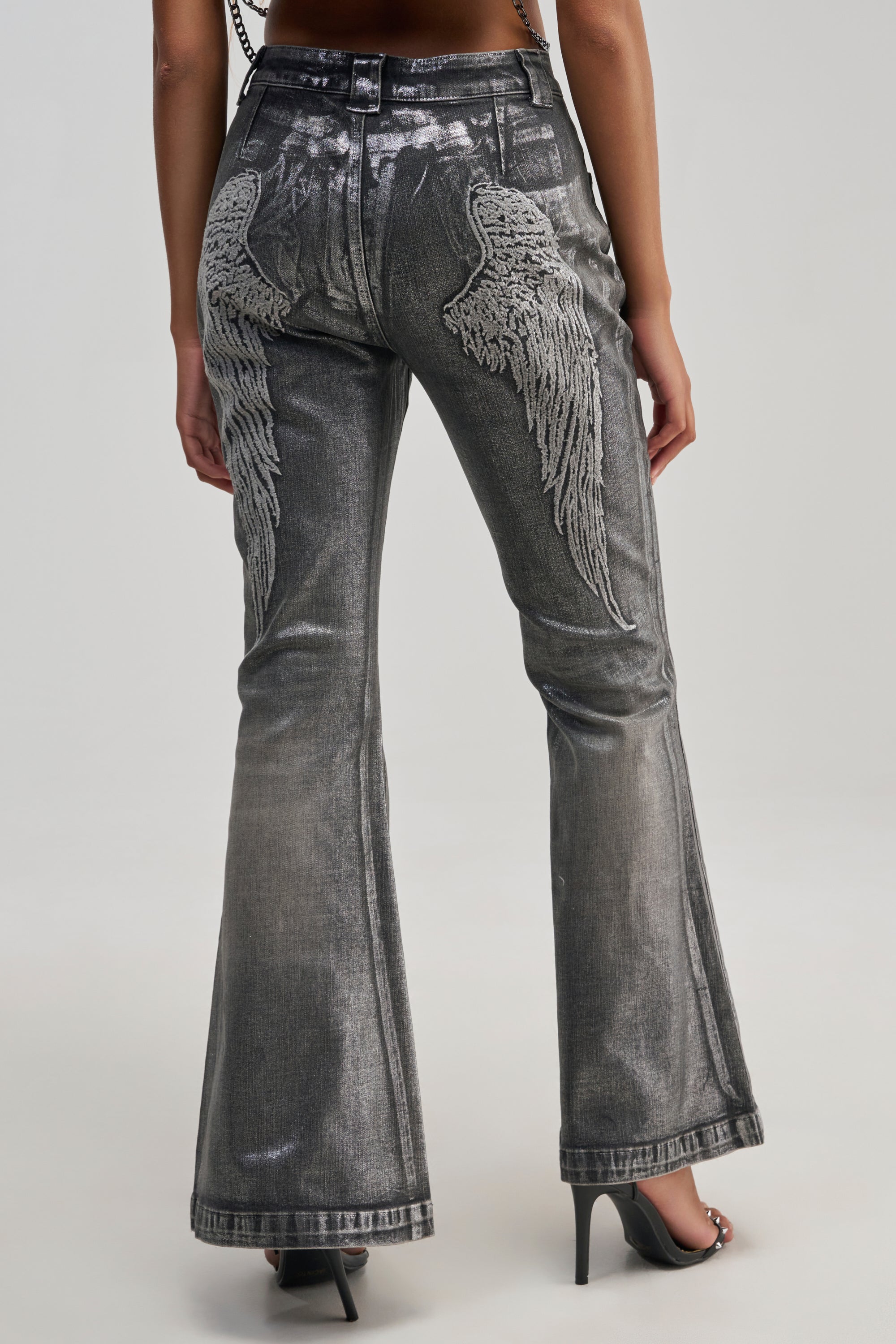 A person is seen from the back wearing THE REAR VIEW WINGS DENIM—silver metallic flared jeans featuring embroidered angel wings on the back pockets—styled with high-heeled shoes for a bold streetwear look.