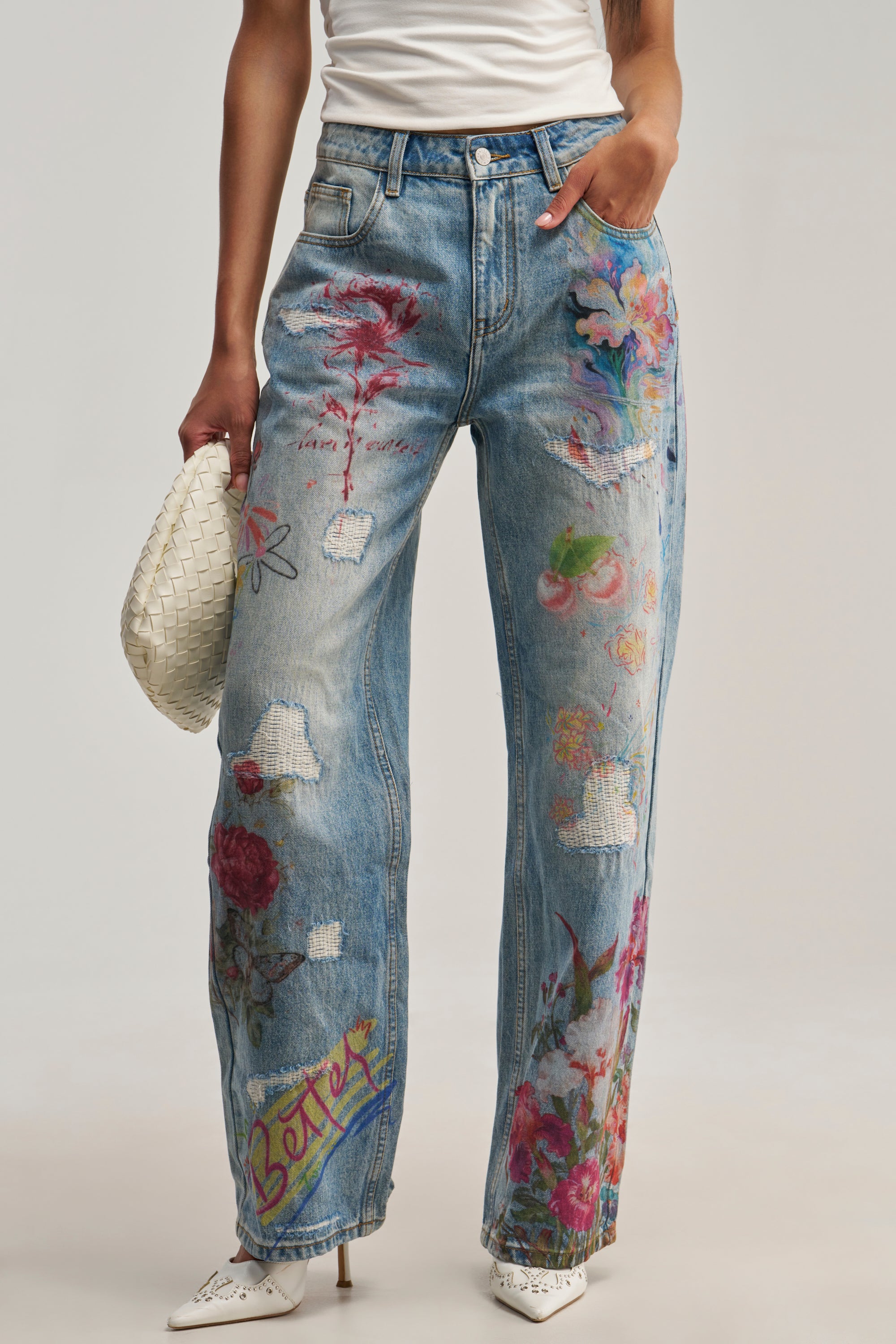 A person wearing BUY ME FLOWERS PRINTED DENIM, featuring multicolor floral and abstract designs, paired with a white woven clutch bag and white pointed shoes. Only the lower body is visible.