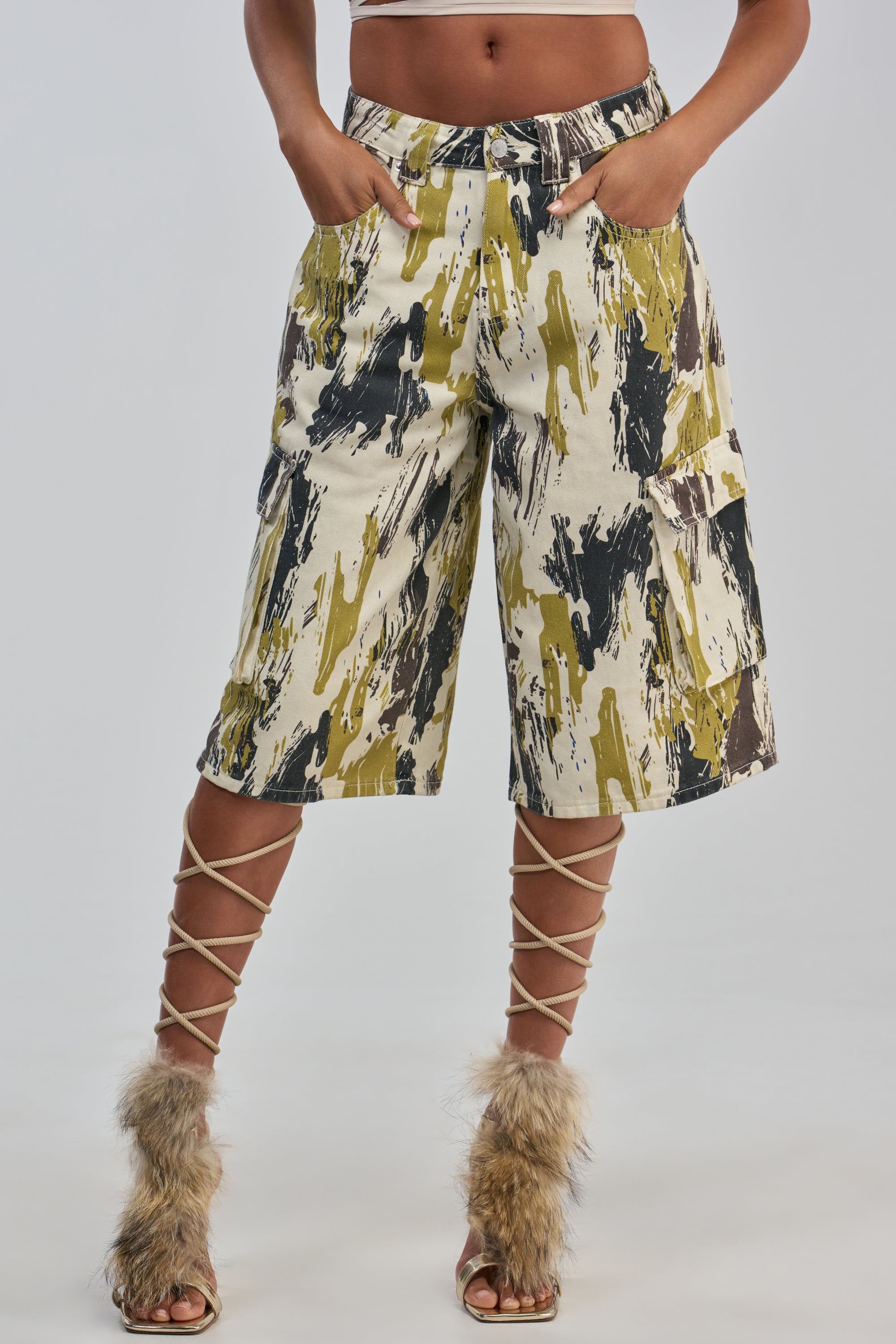 A woman wears the IS THIS CRAZY ENOUGH BERMUDA SHORT, featuring a knee-length silhouette, abstract earth-tone print, and cargo-style pockets, with her hands casually tucked in. She completes the look with lace-up sandals adorned with faux fur at the toes for a hint of streetwear flair. The background is plain and light-colored.