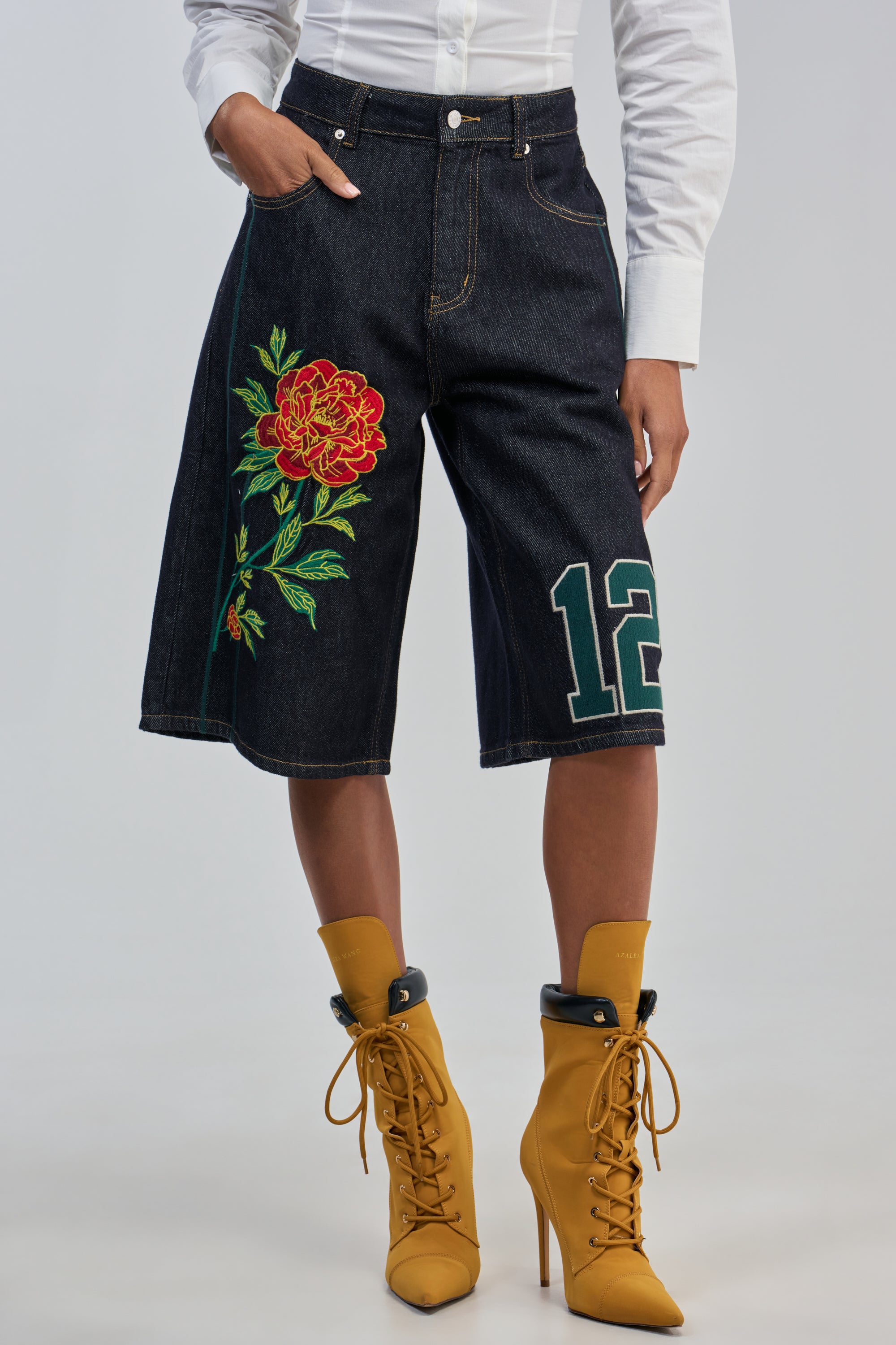 Model sporting the GAME OVER ROSE DENIM BERMUDA SHORTS—knee-length, dark denim shorts featuring striking embroidered patches like a vibrant red rose and a green “12”—teamed with a crisp white shirt and mustard yellow lace-up heeled boots, photographed against a plain backdrop for an effortlessly cool streetwear look.