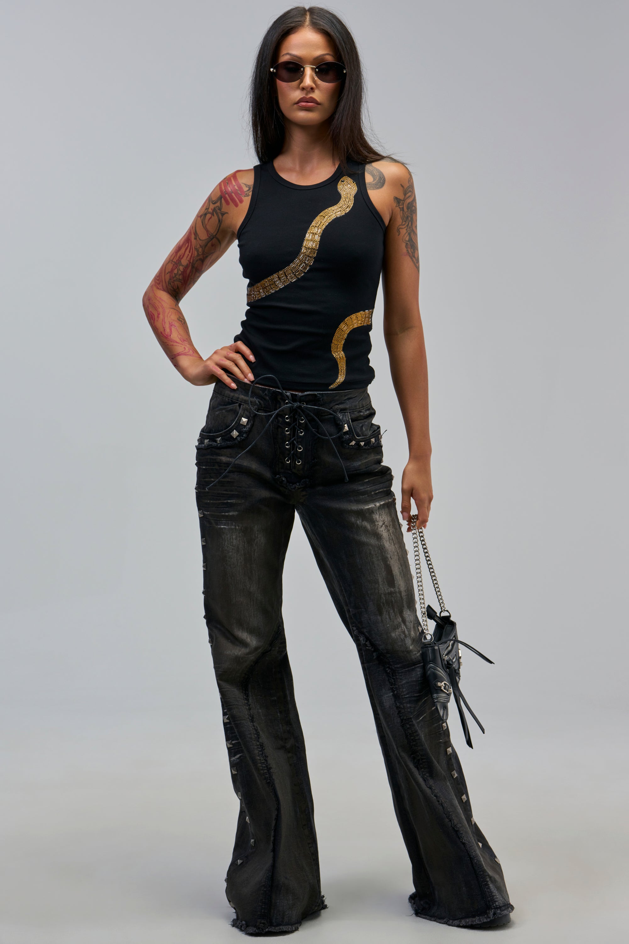 A woman with long dark hair and sunglasses stands confidently, wearing CRAZY FOR YOU METALLIC DENIM, a black tank with a gold snake design, and holding a small black handbag to showcase her fall streetwear and tattoos.
