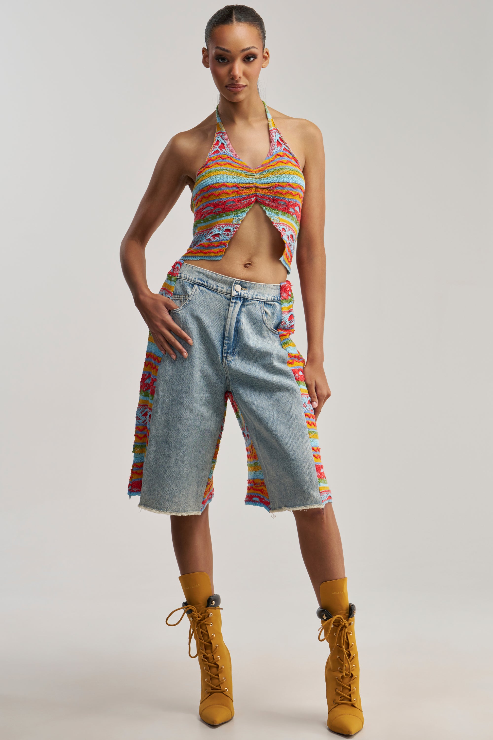 A person poses in an abstract knit multicolored halter top, the FEELS LIKE SUMMER KNIT BERMUDA SHORT, and yellow lace-up boots with matching socks against a plain background.