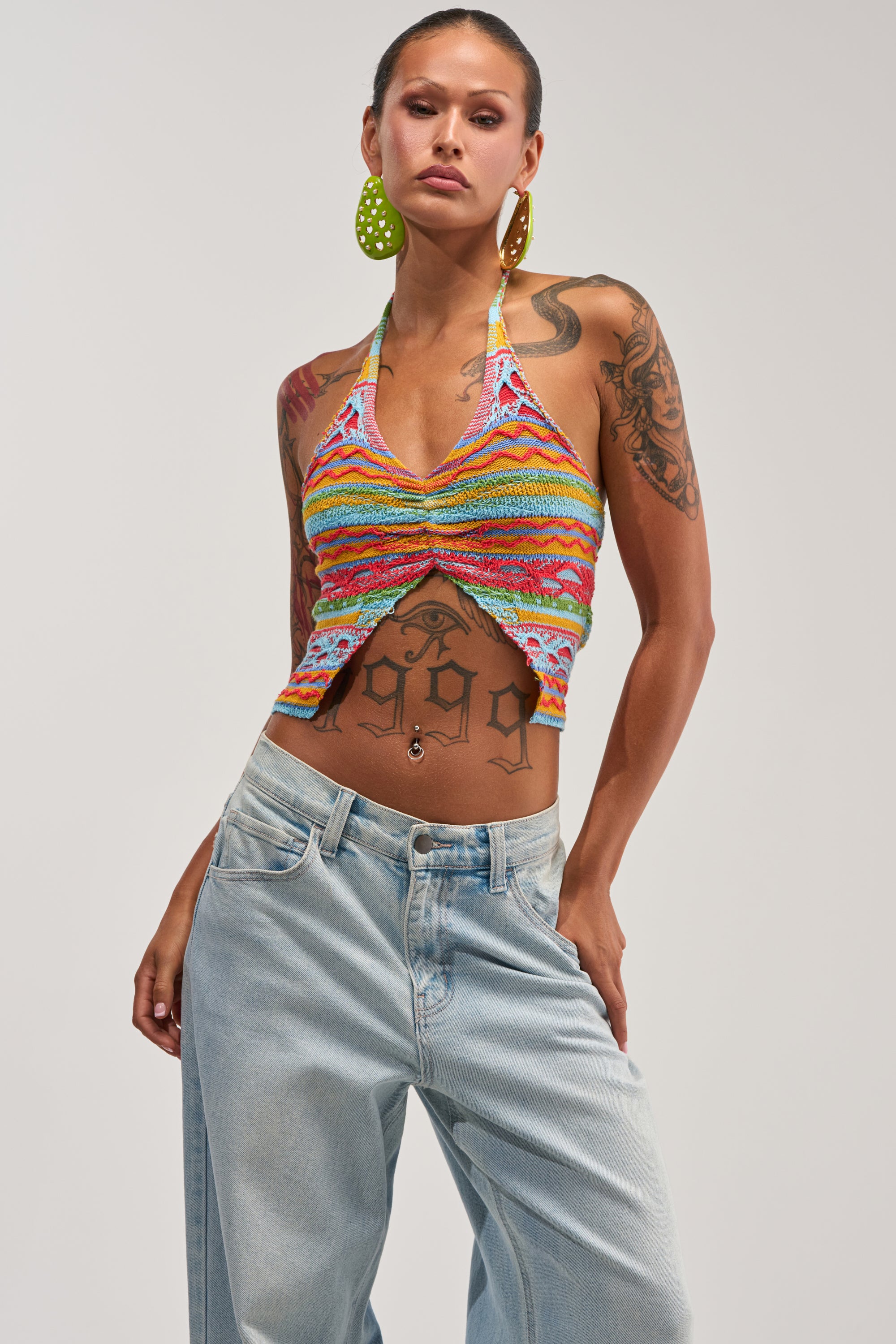 Wearing the FEELS LIKE SUMMER KNIT HALTER TOP—a colorful striped crochet halter—paired with light blue jeans and large yellow earrings, a confident individual with tattoos on their arms and torso poses with their hair pulled back.