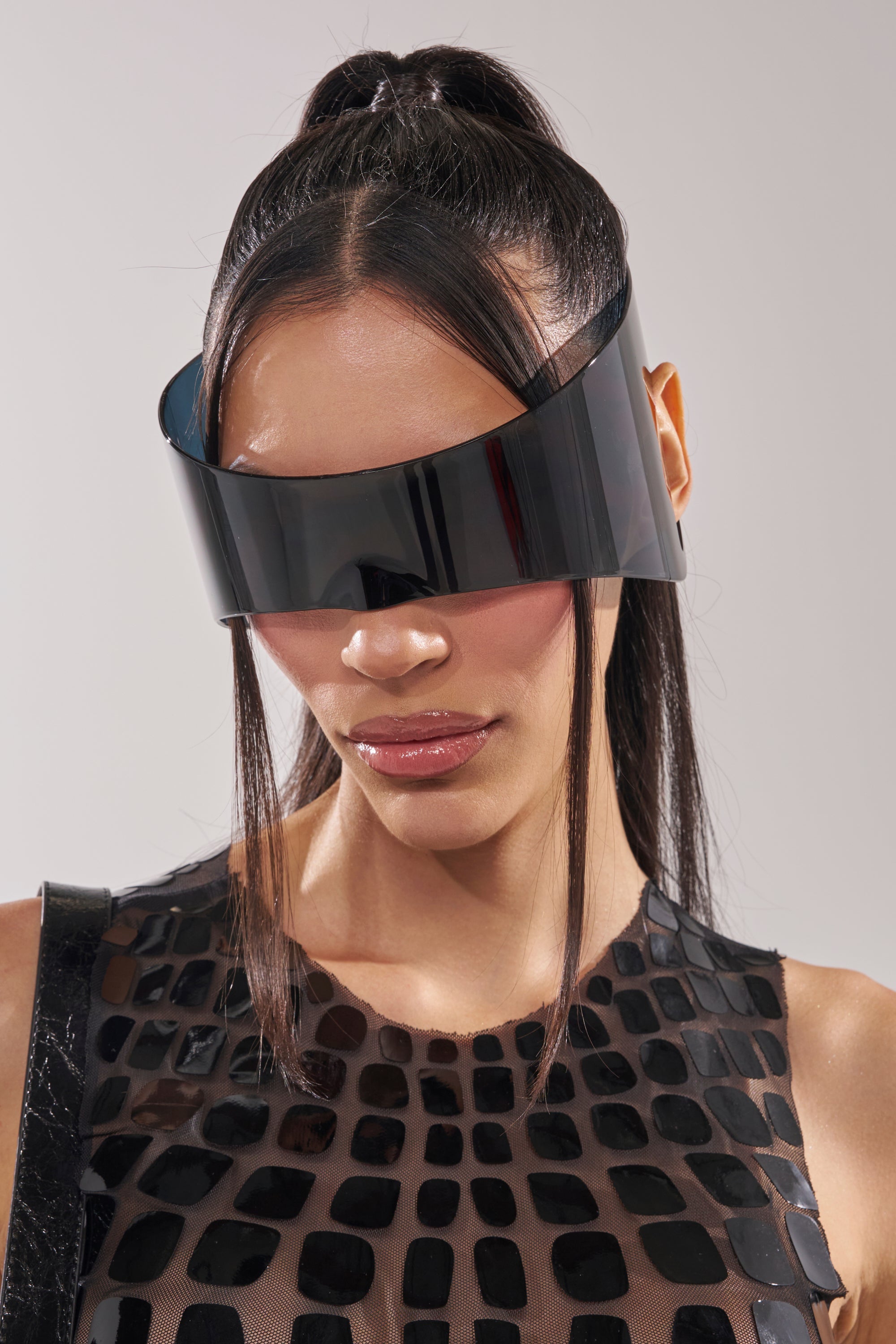 A woman with straight dark hair in a high ponytail wears IRVY SUNGLASSES—large, futuristic black shield frames—and a black, textured rectangular-patterned top against a neutral background. 1