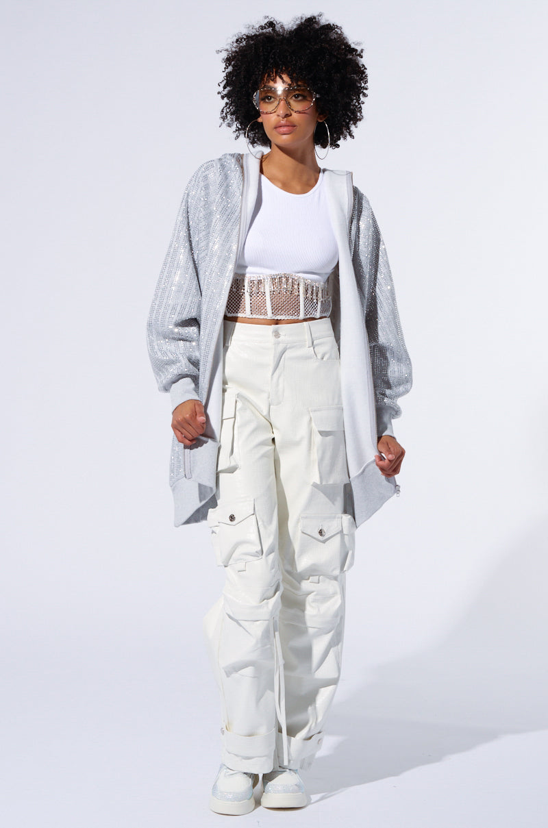 A person with curly hair is wearing the TRES CHIC PASSPORT OVERSIZE ZIP UP JACKET in light gray over a white crop top, paired with beige cargo pants and white sneakers. They have hoop earrings and tinted glasses, posing against a plain background.