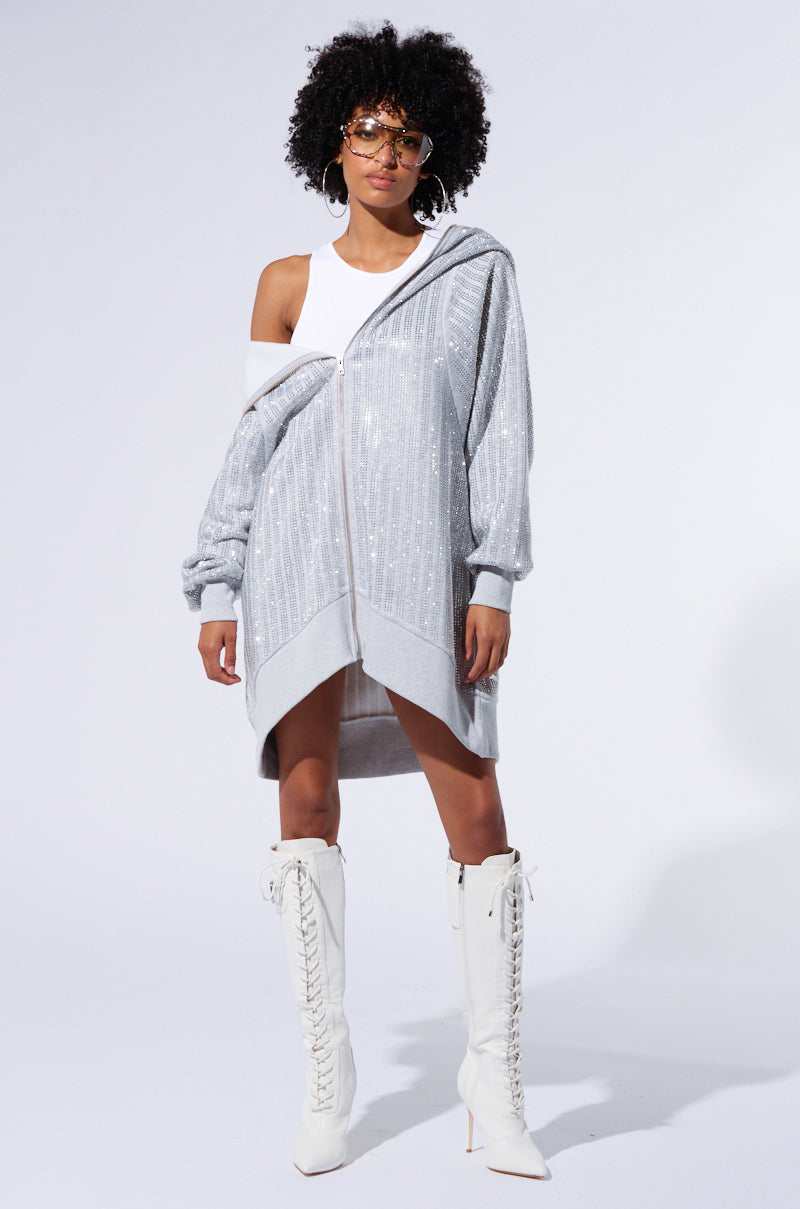 A person with curly hair wearing large glasses, a white tank top, a long, silver TRES CHIC PASSPORT OVERSIZE ZIP UP JACKET with rhinestone embellishments, and white knee-high lace-up boots stands against a plain background.
