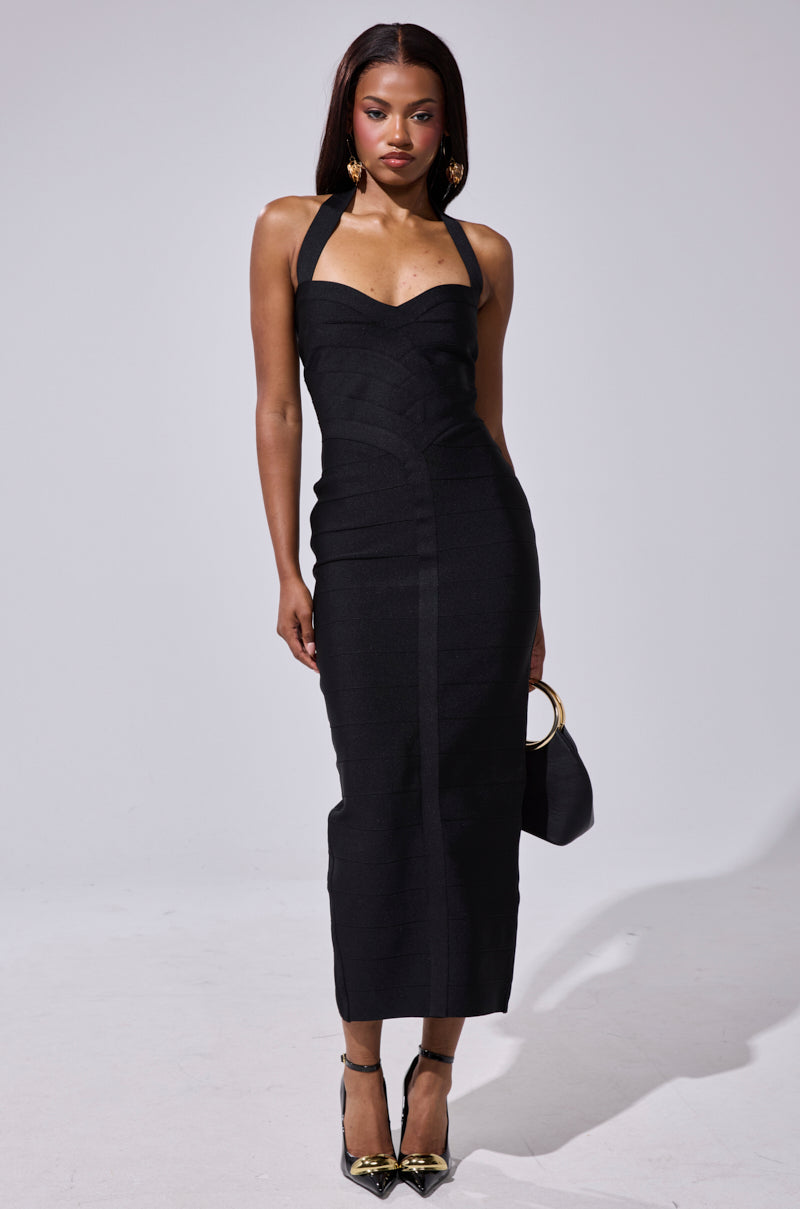 A woman stands against a plain background wearing the LOVELY DAYS BANDAGE MIDI DRESS, a fitted strapless black midi dress with halter straps. She holds a small black purse with a gold handle, wears black heels with gold tips, and has straight, loose hair.