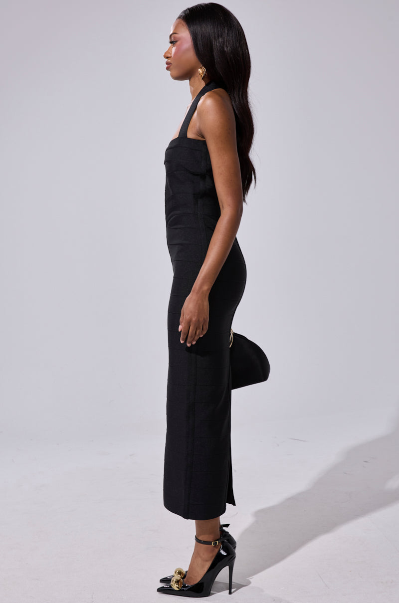 A woman with long dark hair stands in profile against a plain light background, wearing the LOVELY DAYS BANDAGE MIDI DRESS, black heels with gold accents, gold earrings, and holding a black clutch bag.