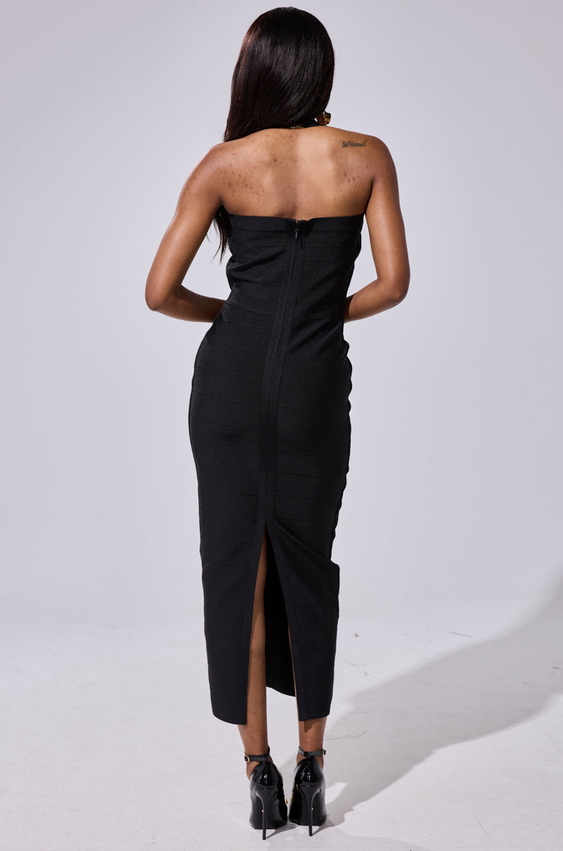 A woman stands facing away in the LOVELY DAYS BANDAGE MIDI DRESS, a strapless fitted black midi with back slit and zipper. Her long dark hair is down, and she wears black heeled shoes.
