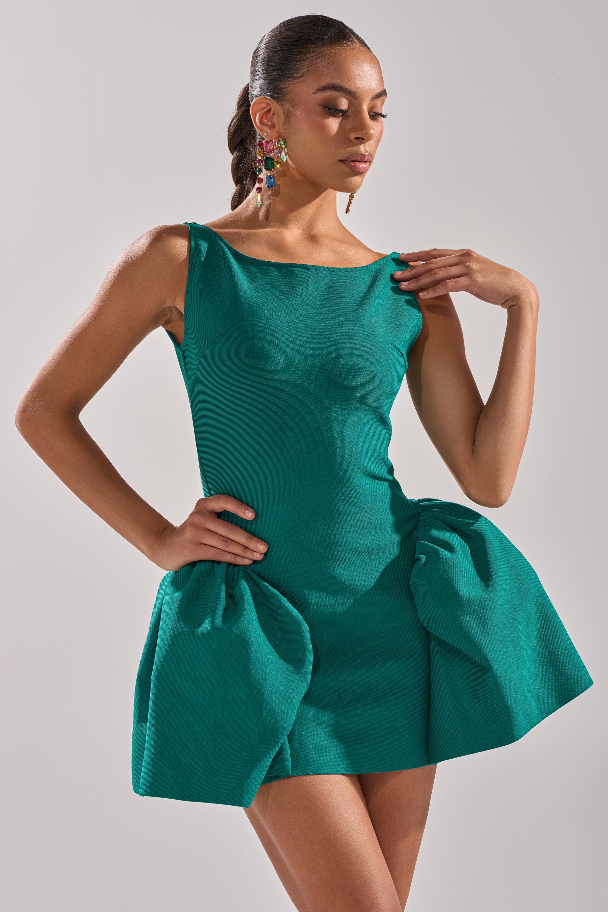 A woman models the DERBY DAYS BANDAGE MINI DRESS, a sleeveless green party dress with a sculpted bodice and dramatic gathered hip details. She pairs it with colorful statement earrings and wears her hair in a sleek low bun. 1