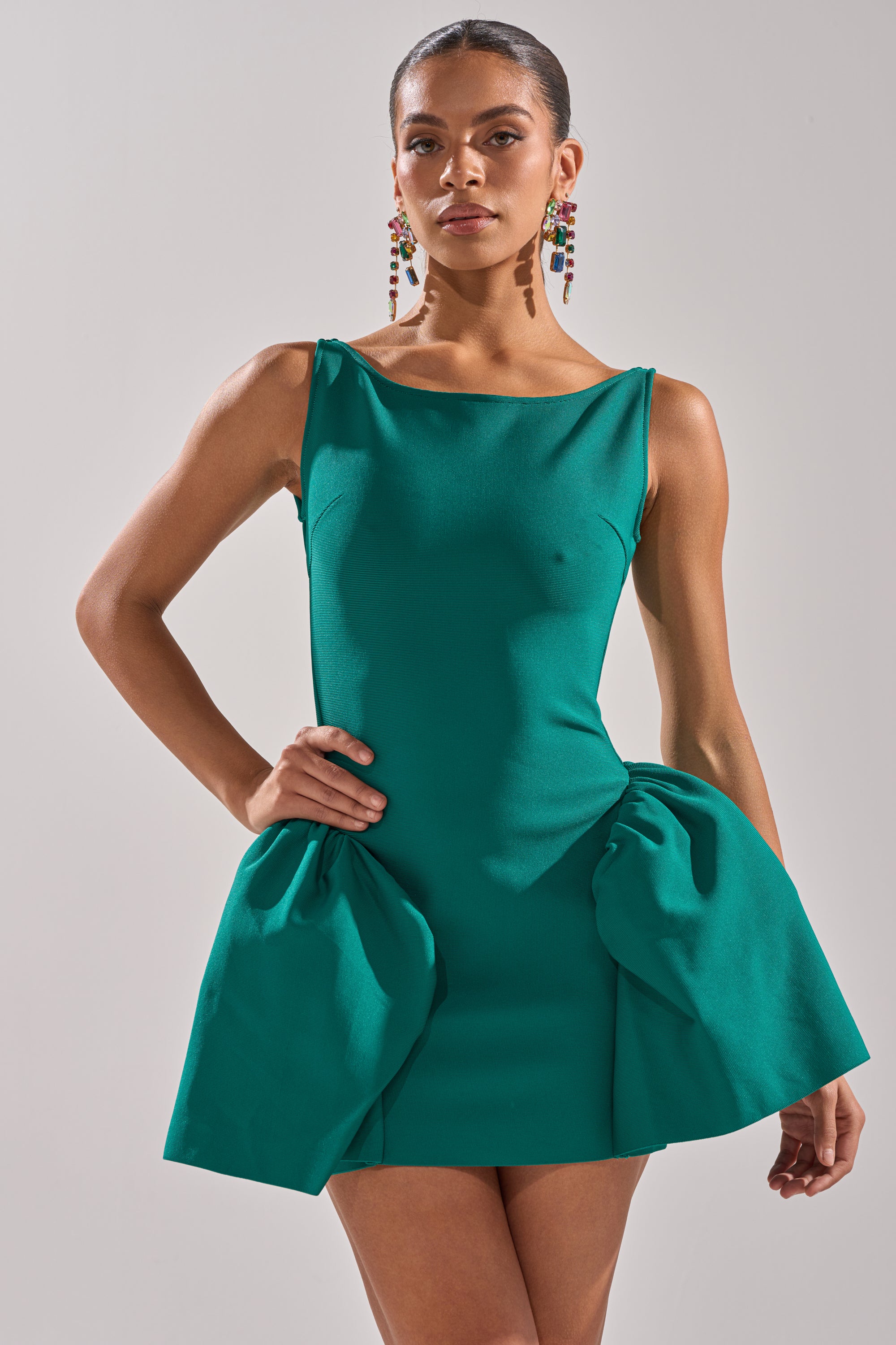 Woman wearing the DERBY DAYS BANDAGE MINI DRESS—sleeveless, emerald green, with a sculpted bodice and gathered hip details—stands against a plain background, one hand on her hip, accessorized with colorful beaded earrings. 2