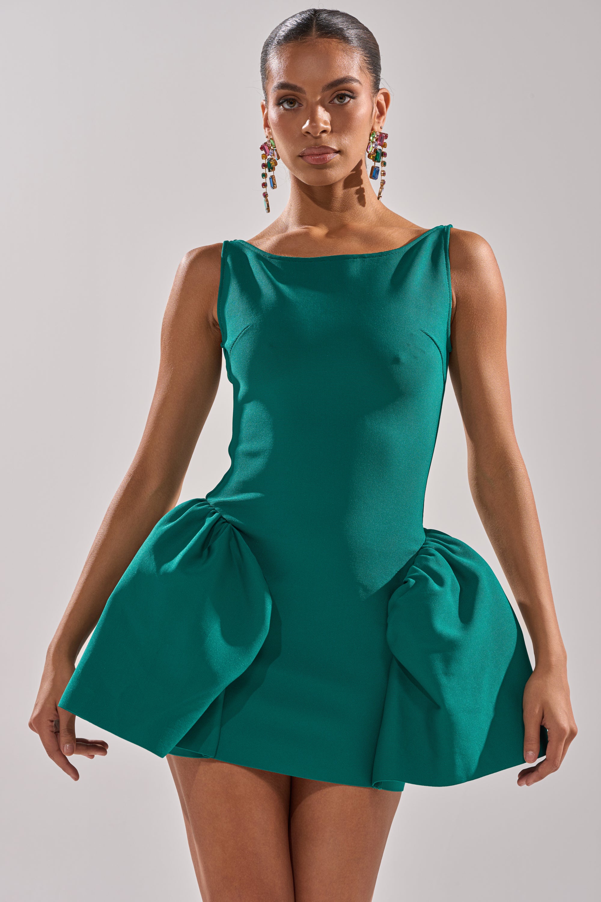 A woman wears the DERBY DAYS BANDAGE MINI DRESS in emerald green, featuring sleeveless styling and sculpted hip ruffles. She stands against a light background with sleek pulled-back hair and long, colorful earrings. 3