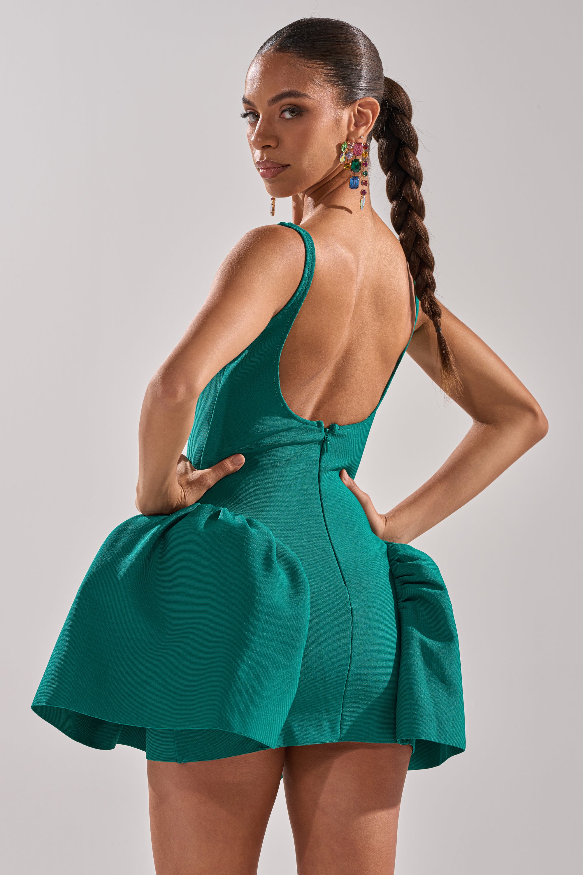 A woman with a long braided ponytail wears the DERBY DAYS BANDAGE MINI DRESS, a sleeveless, backless green party dress with voluminous side ruffles. She stands confidently against a plain background, hands on hips. 5