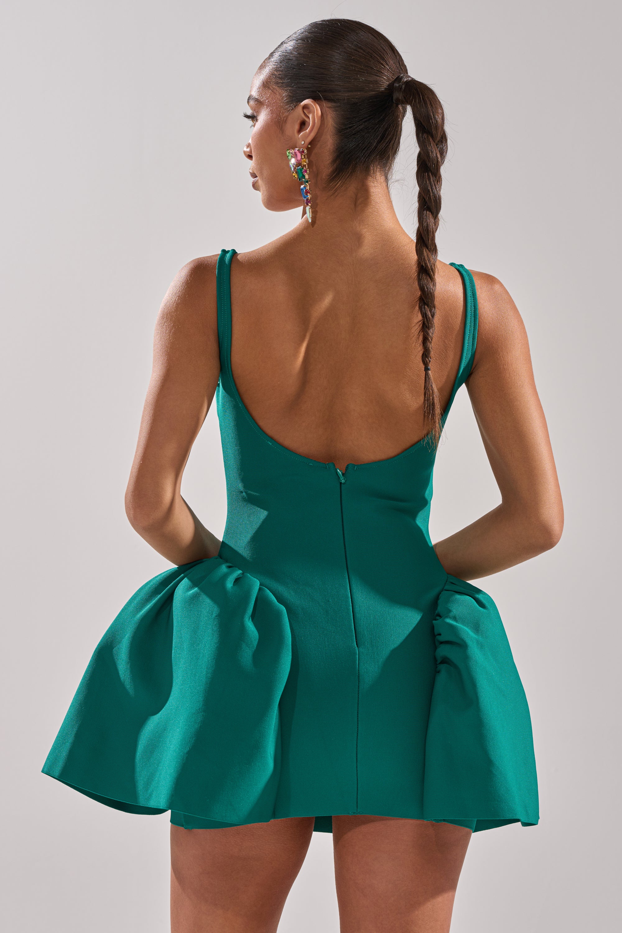 A woman with a braided ponytail stands with her back to the camera, wearing the sleeveless green DERBY DAYS BANDAGE MINI DRESS with a flared skirt and colorful earrings. Her hands rest on her hips against a neutral backdrop. 6
