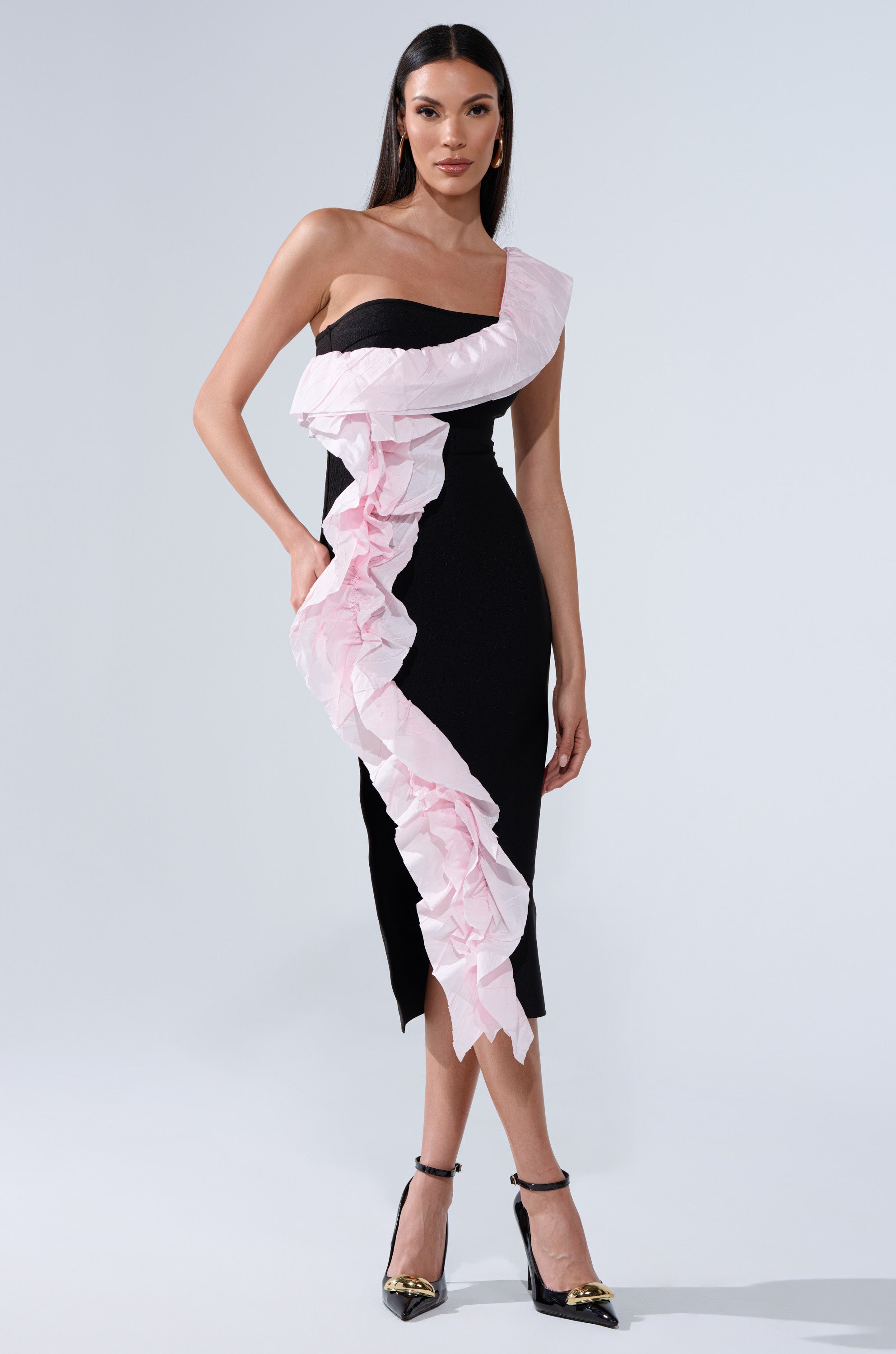 She wears the SOFT FLEX BANDAGE MIDI DRESS WITH RUFFLE DETAIL, a striking black one-shoulder dress with a bold pink ruffle spiraling from shoulder to hem. Her look is finished with sleek, straight hair and black ankle-strap heels.
