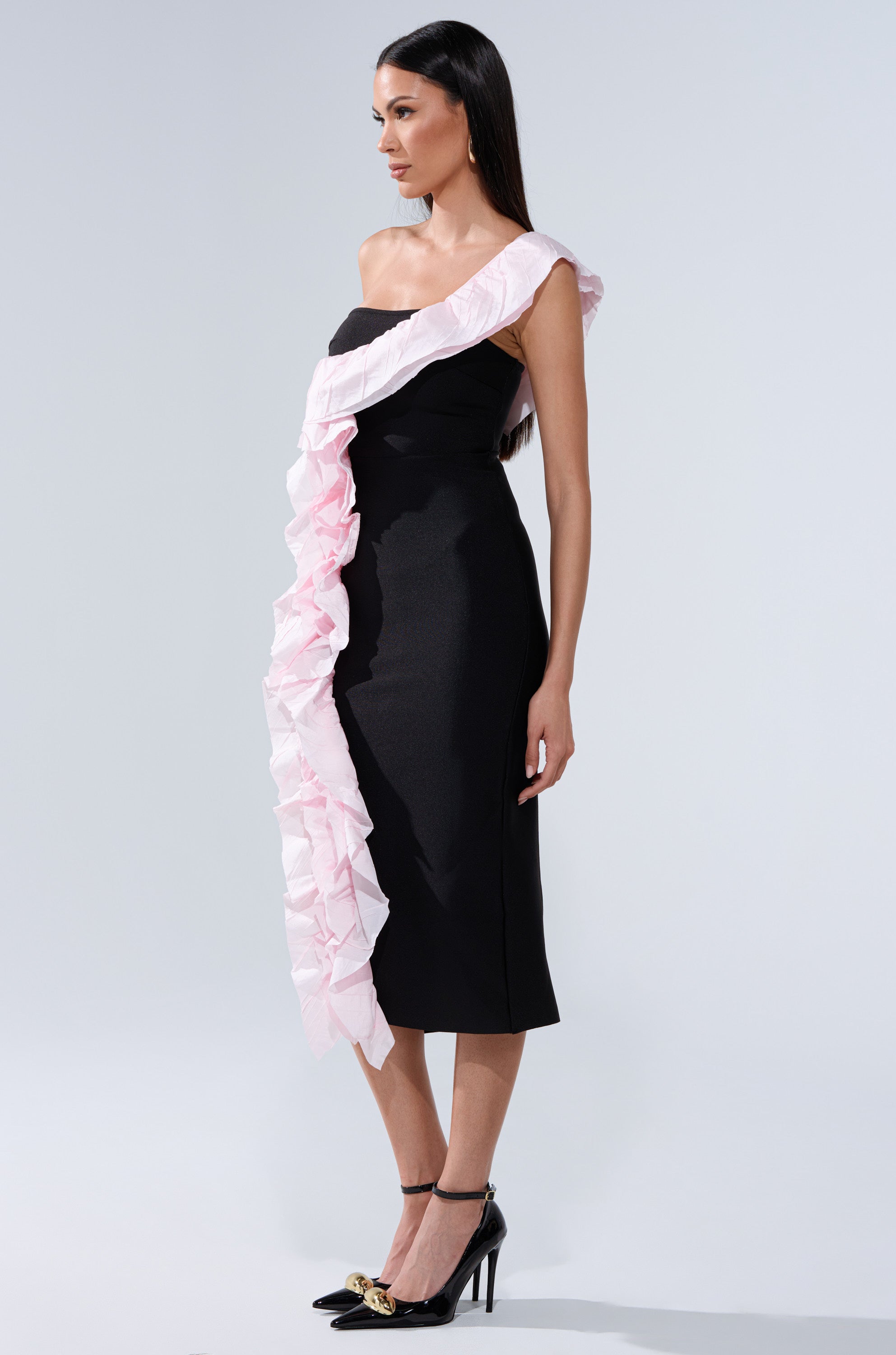 A woman wears the SOFT FLEX BANDAGE MIDI DRESS WITH RUFFLE DETAIL—black, fitted, midi-length with a bold pink ruffle from shoulder to hem—paired with black heels against a plain light background.