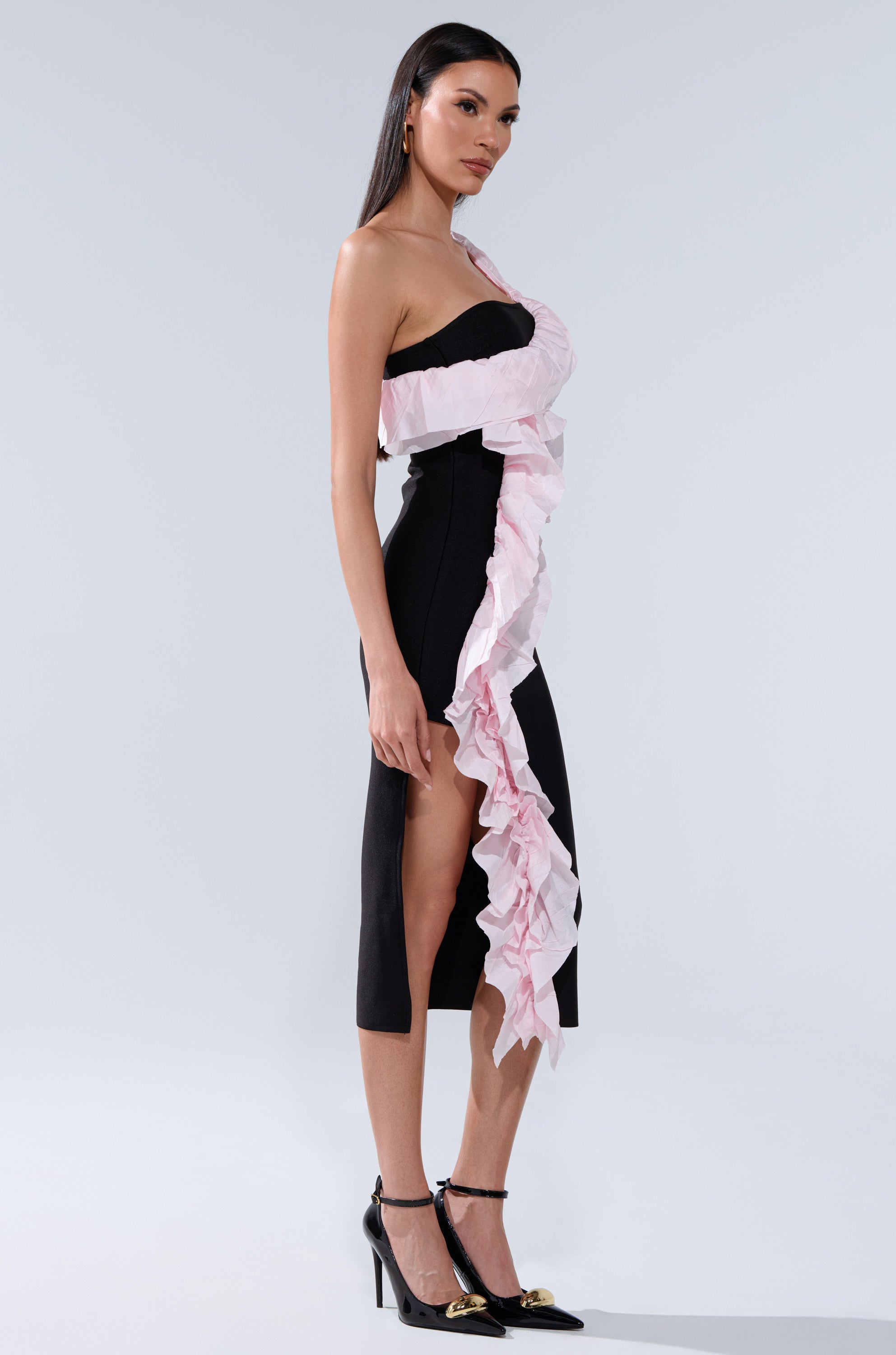 A woman in profile wears the SOFT FLEX BANDAGE MIDI DRESS WITH RUFFLE DETAIL, a black one-shoulder dress featuring ruffles, a high slit, and pink trim. She has long straight hair and completes the look with black heels.