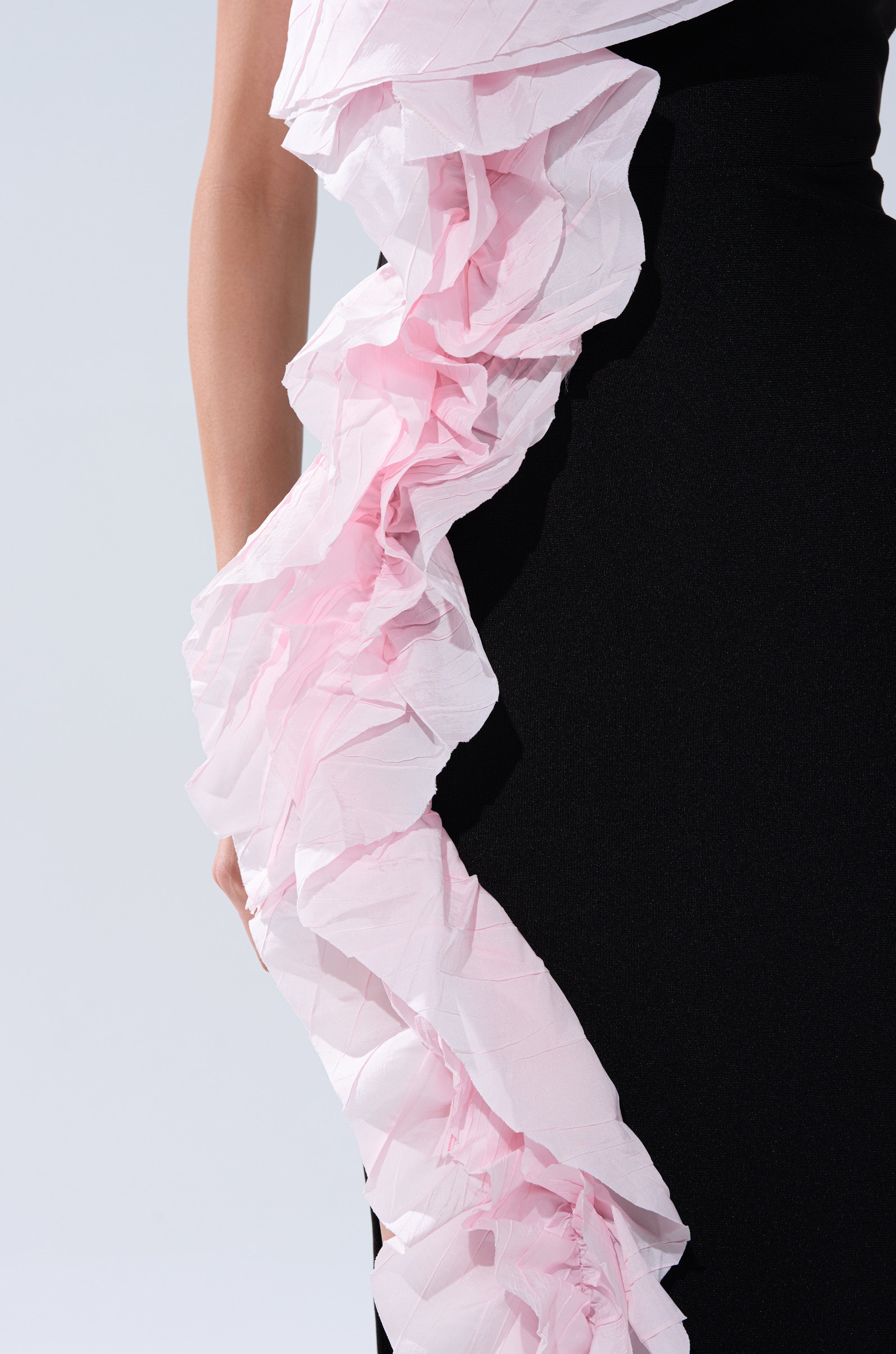 Someone wearing the SOFT FLEX BANDAGE MIDI DRESS WITH RUFFLE DETAIL, a black midi dress featuring a large light pink ruffle running diagonally along the side, stands against a plain light background.