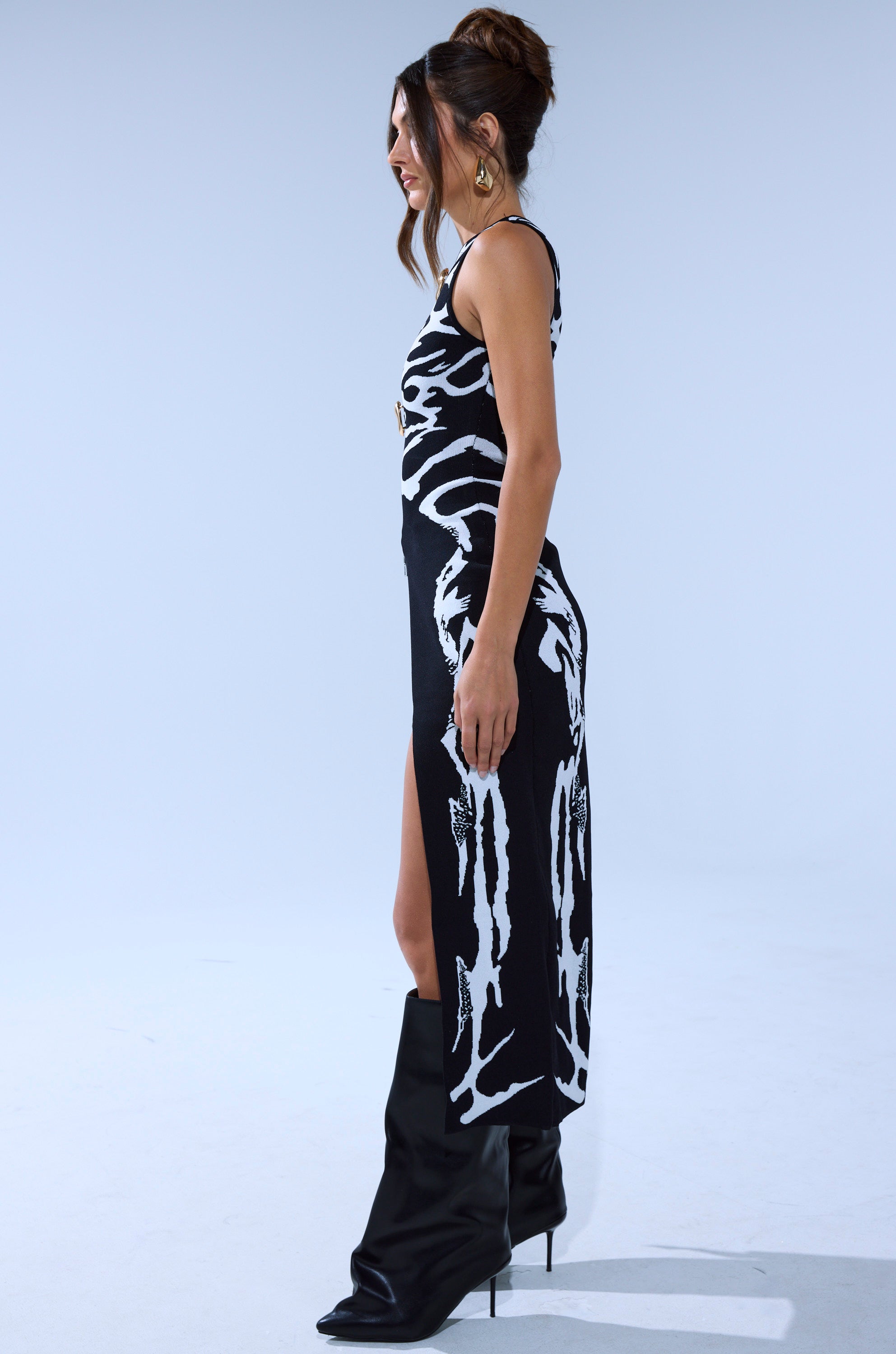 A woman stands sideways in the INCOGNITO SWEATER, a sleeveless abstract black and white dress with a thigh-high slit, paired with black knee-high boots. Her hair is styled in a bun and she wears large hoop earrings. 4