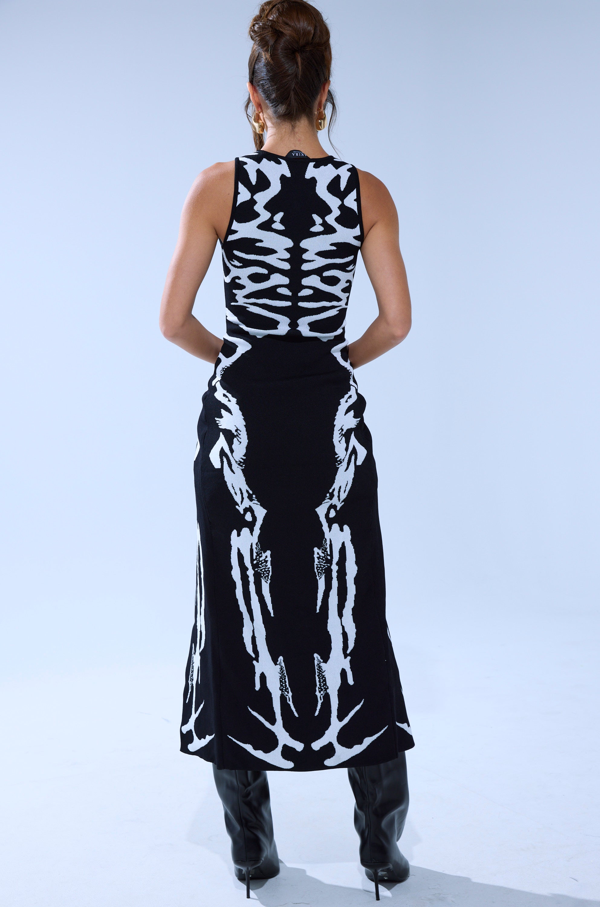 A woman with an updo wears the INCOGNITO SWEATER, a sleeveless black dress with bold white abstract back patterns, paired with tall black boots, standing against a plain light background. 5