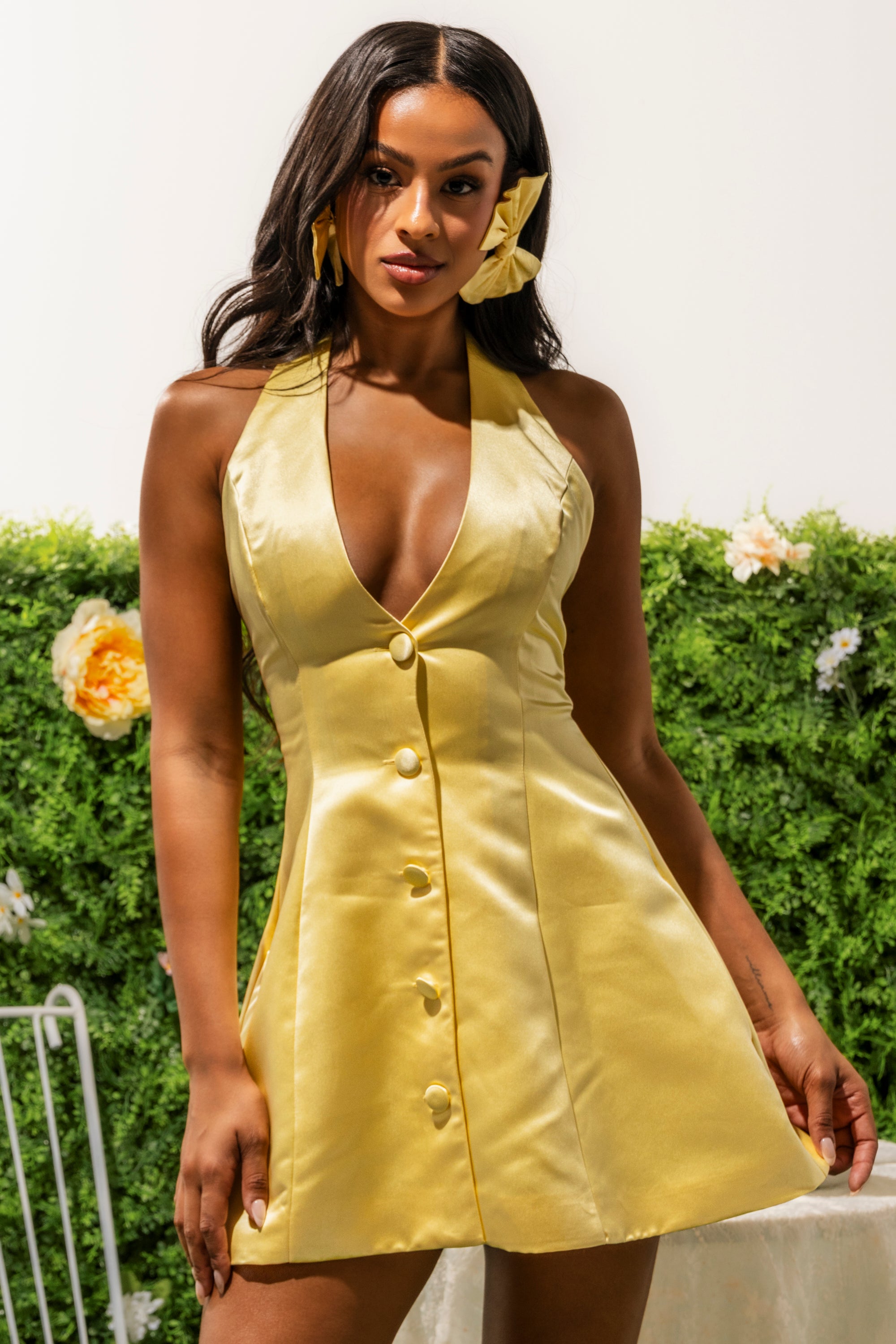 A woman models the SLAY READY BUTTON UP MINI DRESS, a yellow halter mini with a deep V-neckline and bold buttons, posing confidently against a green floral backdrop. 1