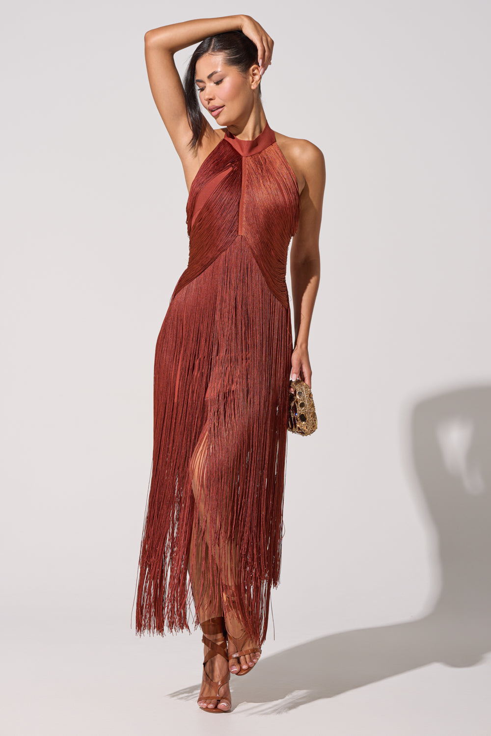 A woman models the FORGET ME NOT FRINGE MAXI DRESS in rust, pairing it with a small clutch and strappy heels. She poses against a plain light background with one arm raised—an ideal look for an evening event. 3