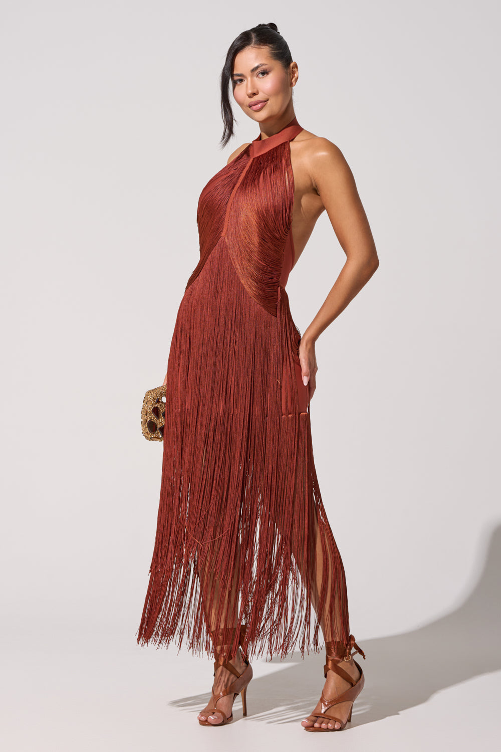 A woman poses in the FORGET ME NOT FRINGE MAXI DRESS, a rust halter-neck maxi with fringe and an asymmetrical hem, styled with brown strappy heels and a small brown bag—an effortlessly chic formal evening look. 4