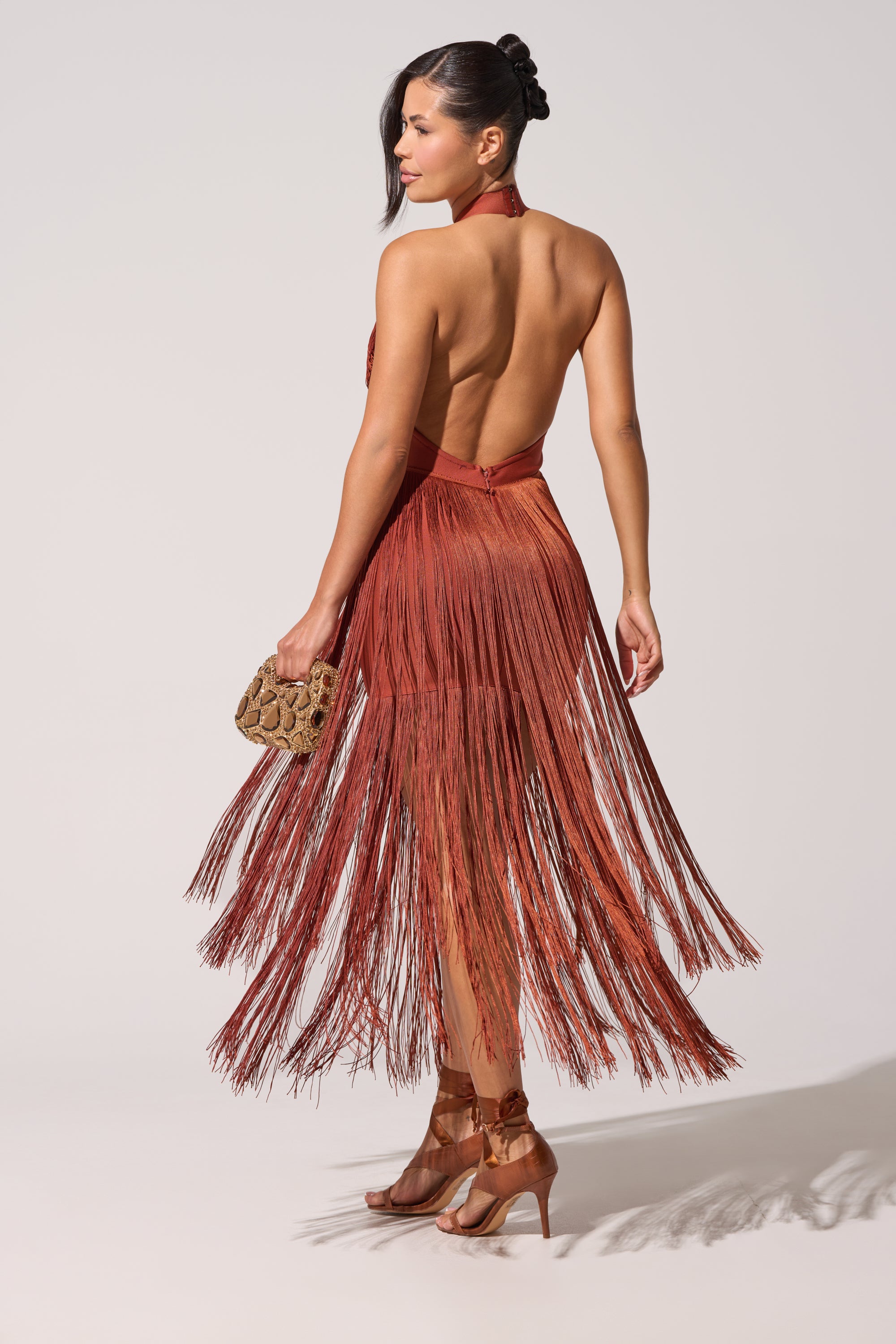 A woman poses against a plain light background wearing the FORGET ME NOT FRINGE MAXI DRESS in rust, styled with brown heels and a small textured clutch. Her hair is sleekly tied back in a ponytail. 1