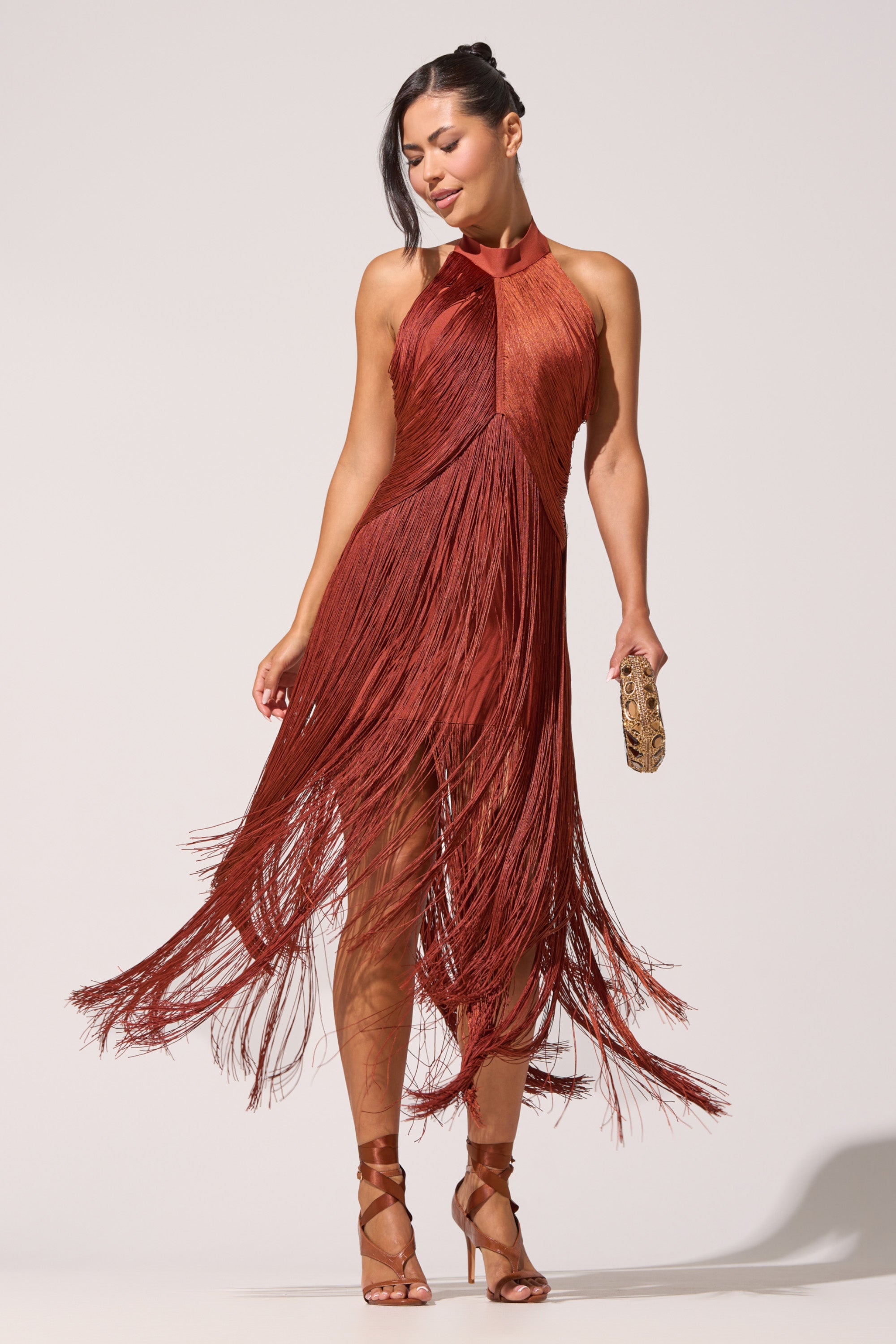 A woman models the FORGET ME NOT FRINGE MAXI DRESS in rust, featuring a halter neckline and swinging fringe. She completes her evening-ready look with strappy heels and a gold clutch, standing poised against a plain backdrop. 2