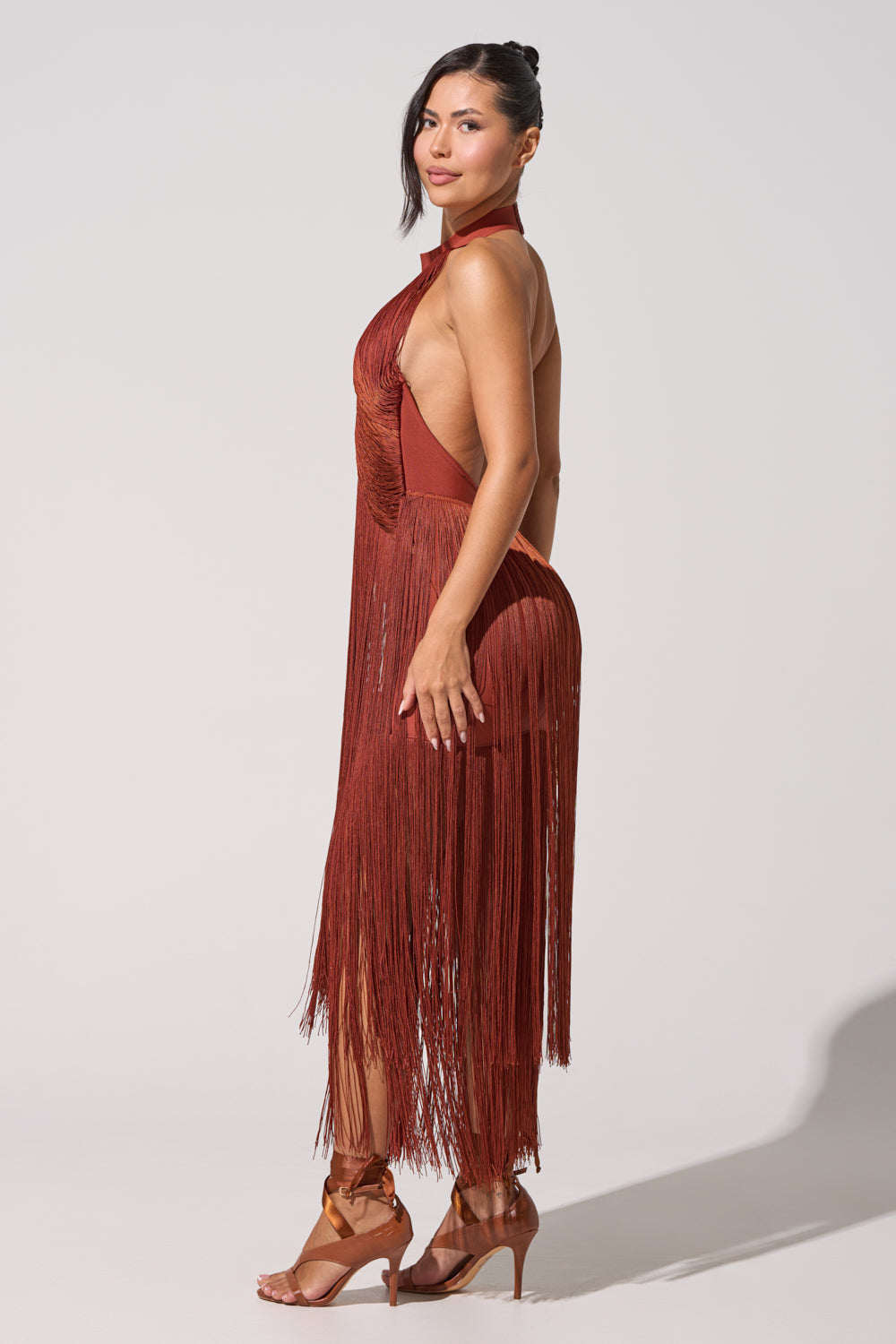 A woman models the FORGET ME NOT FRINGE MAXI DRESS, a rust halter neck sleeveless dress with fringe and open back, styled with brown high-heeled sandals against a plain light background—ideal for evening events. 6