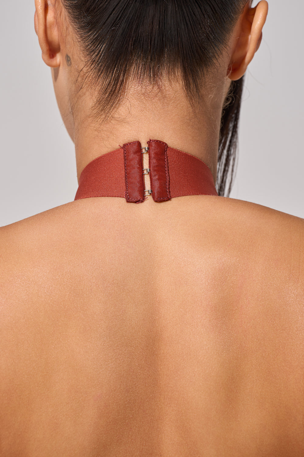 Close-up of a person’s upper back and neck, wearing the FORGET ME NOT FRINGE MAXI DRESS with a maroon choker fastened by metal hooks. Their hair is up, revealing a small tattoo behind the ear—an elegant look for an evening event. 8