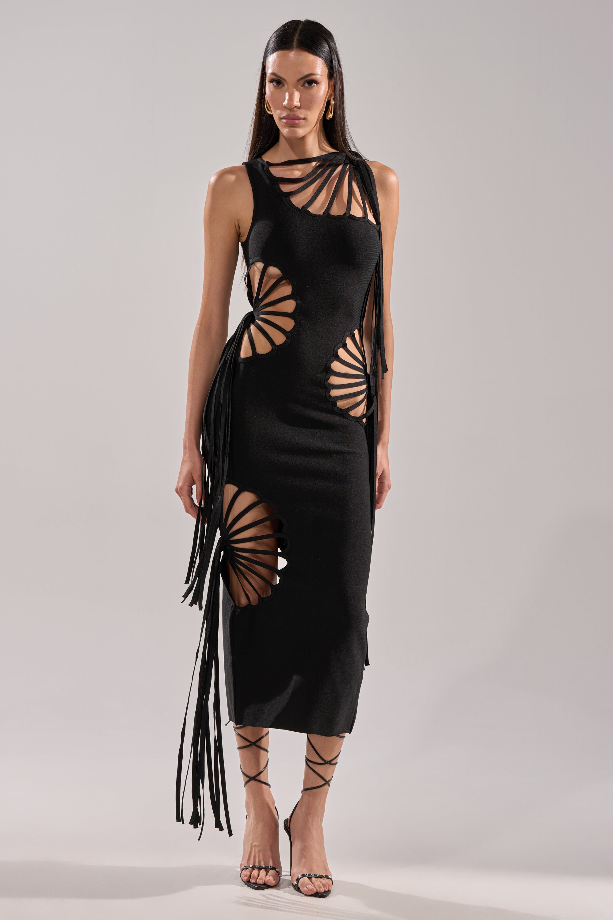 A woman poses against a plain gray background wearing the BROOKE CUTOUT MIDI DRESS, featuring black bandage fabric with large circular cutouts and long fringe, paired with strappy black high-heeled sandals.