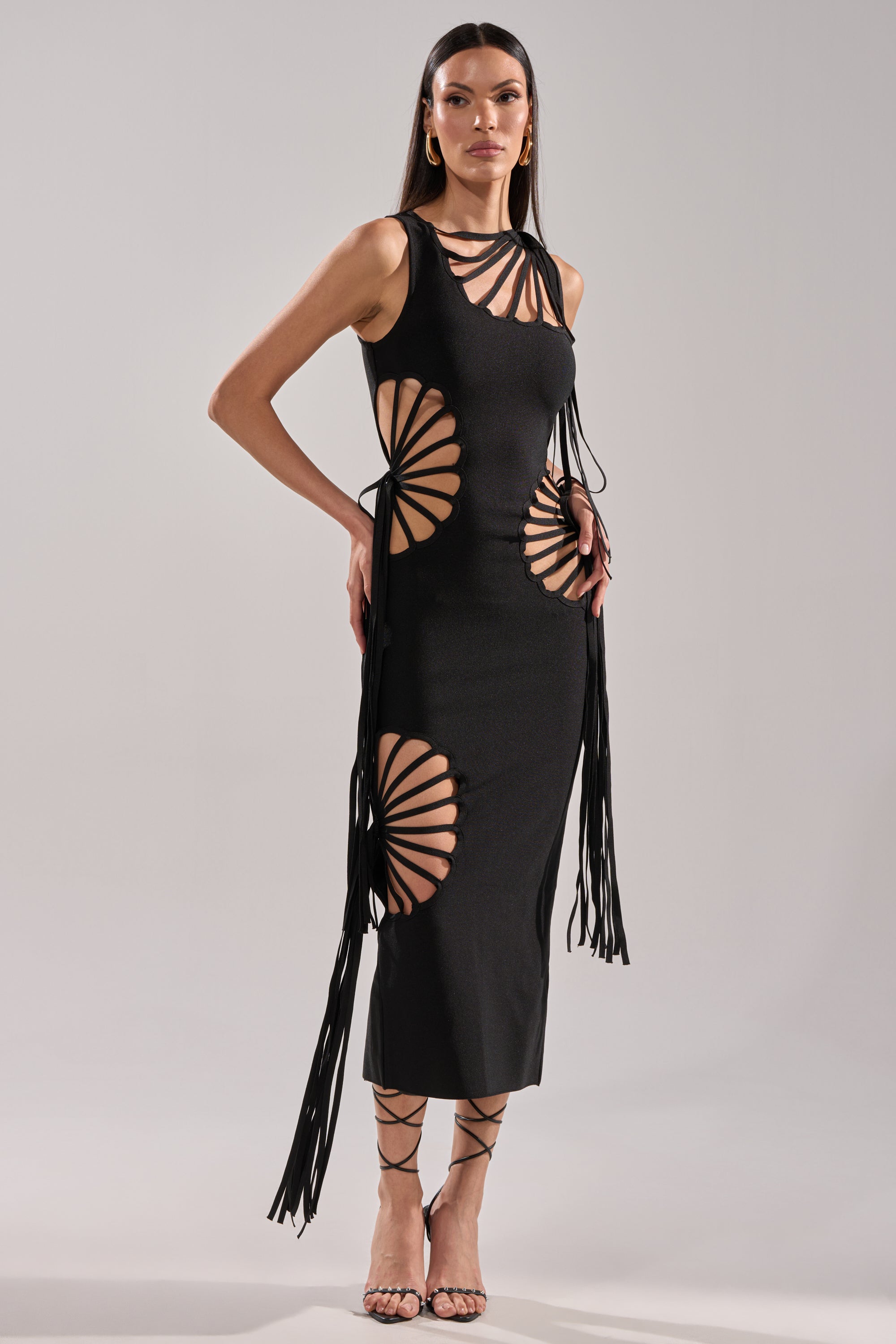 A woman stands against a plain background wearing the BROOKE CUTOUT MIDI DRESS, a sleeveless black bodycon midi featuring dramatic cut-outs, radiating strap details on the sides and chest, long dangling ties, and strappy high-heeled sandals.