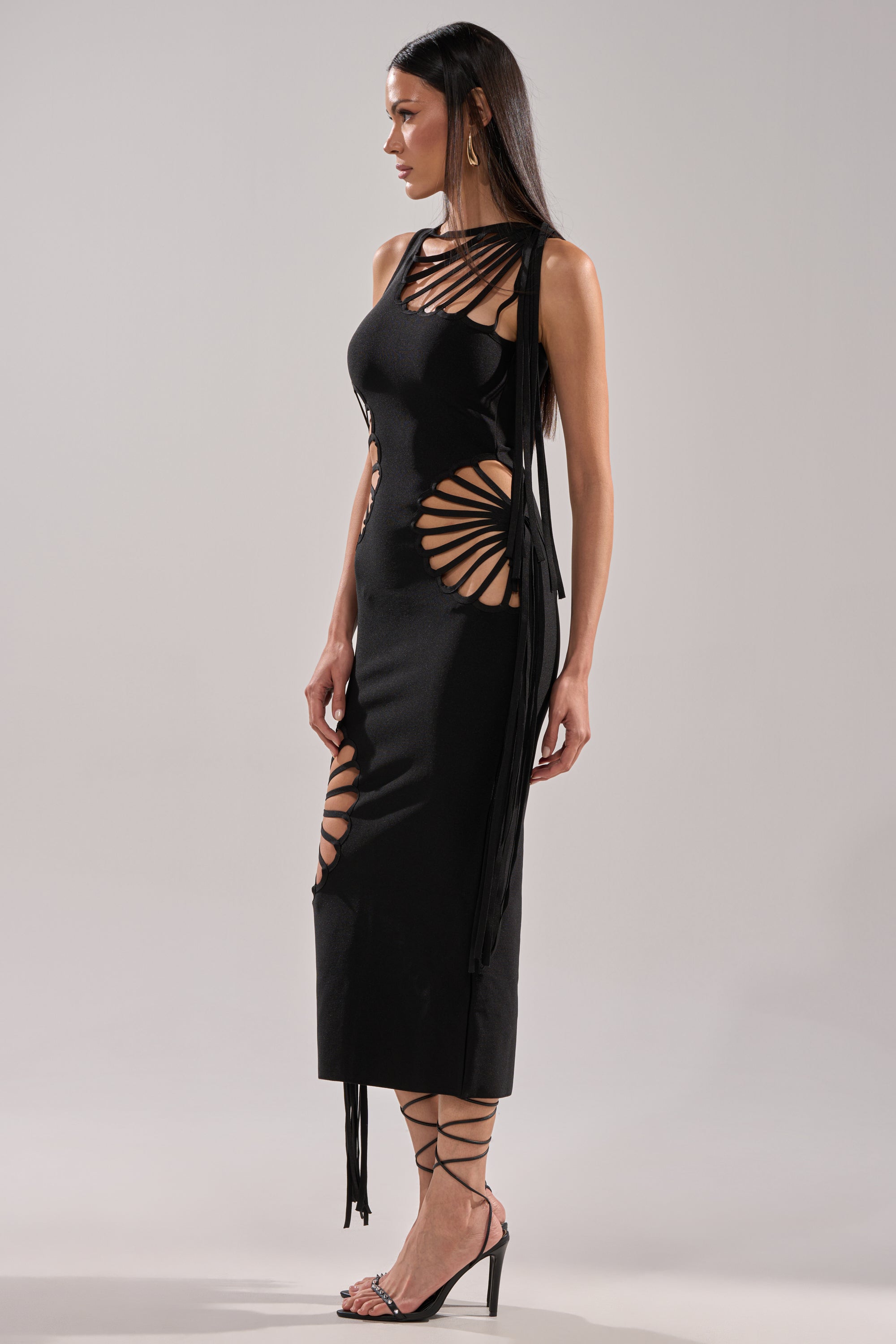 A woman stands in profile wearing the BROOKE CUTOUT MIDI DRESS, a black bodycon style with circular cutouts and radiating straps. She has long straight hair and strappy high heels, set against a plain light background.