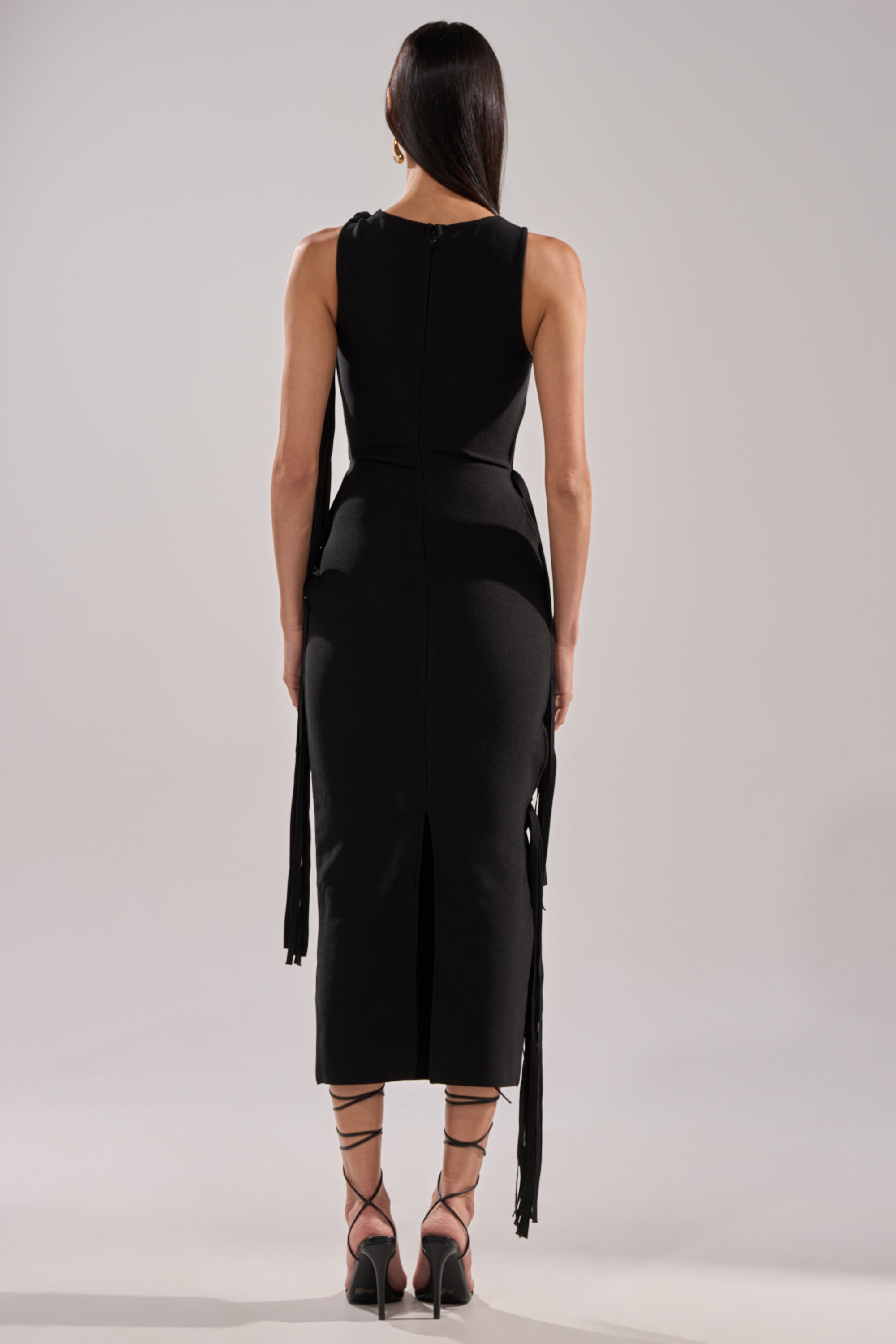 A woman with straight dark hair faces away, modeling the BROOKE CUTOUT MIDI DRESS— a fitted black bodycon style with a back slit—paired with black ankle-tie heels and drop earrings against a plain light background.