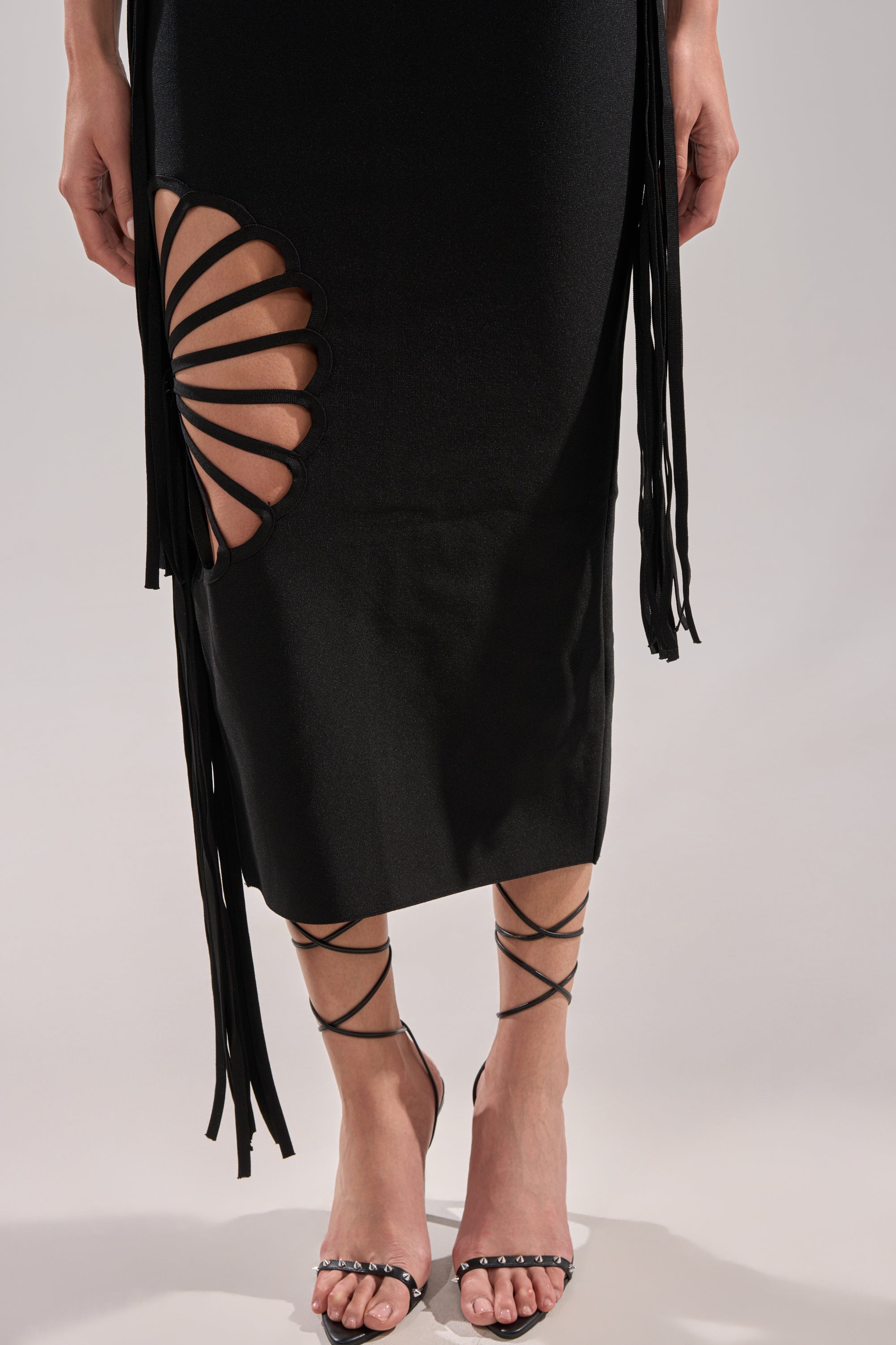 Wearing the BROOKE CUTOUT MIDI DRESS, featuring a black design with a unique thigh pattern and fringe detail, paired with strappy high-heeled sandals that wrap around the ankles.