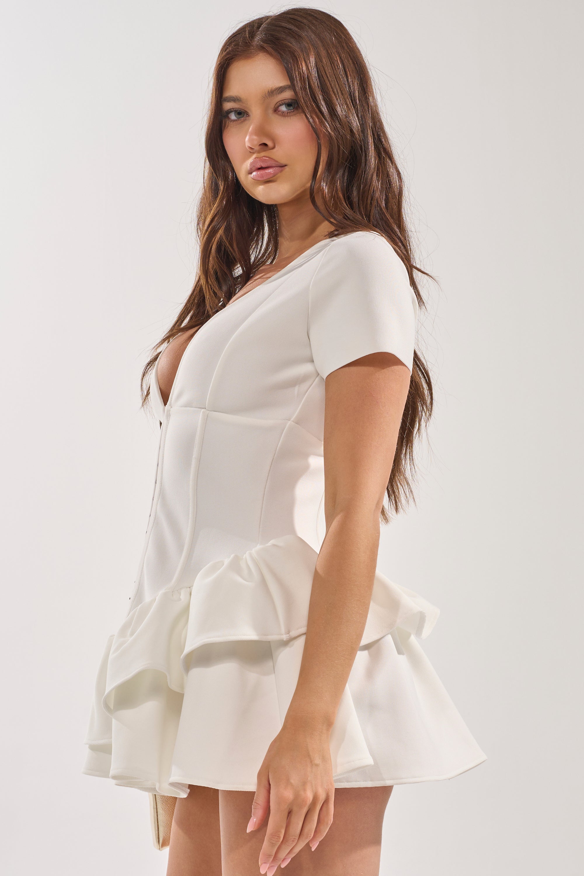 A woman with long brown hair wears the IN BLOOM CORSET RUFFLE DRESS—a white bandage mini dress with a corset-style bodice and ruffled skirt—posing sideways against a plain light background, perfect for bridal events. 2