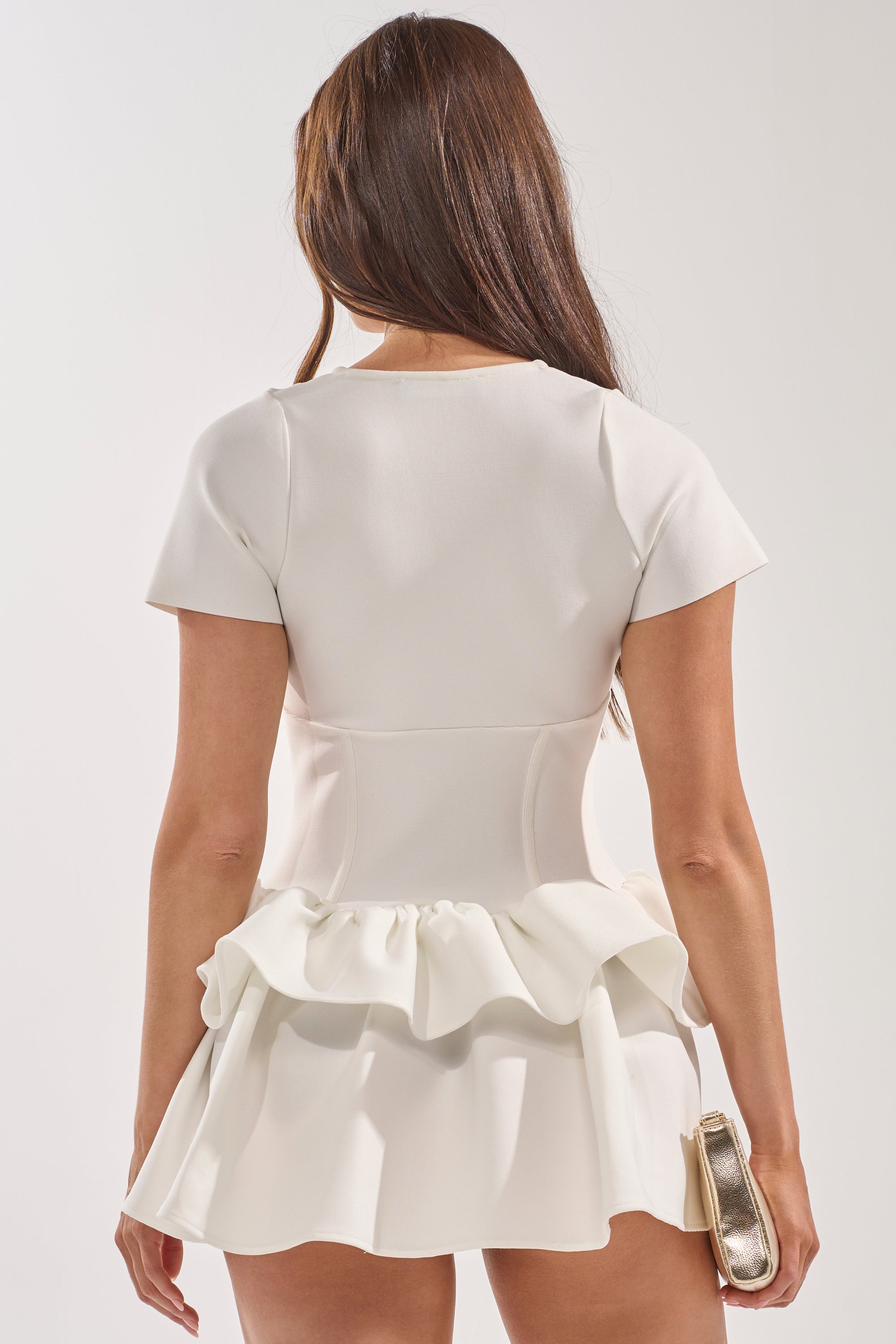 A woman with long brown hair stands facing away, wearing the IN BLOOM CORSET RUFFLE DRESS—a white bandage mini dress with ruffled peplum and flared skirt—perfect for bridal events. She holds a small metallic clutch in her right hand. 3