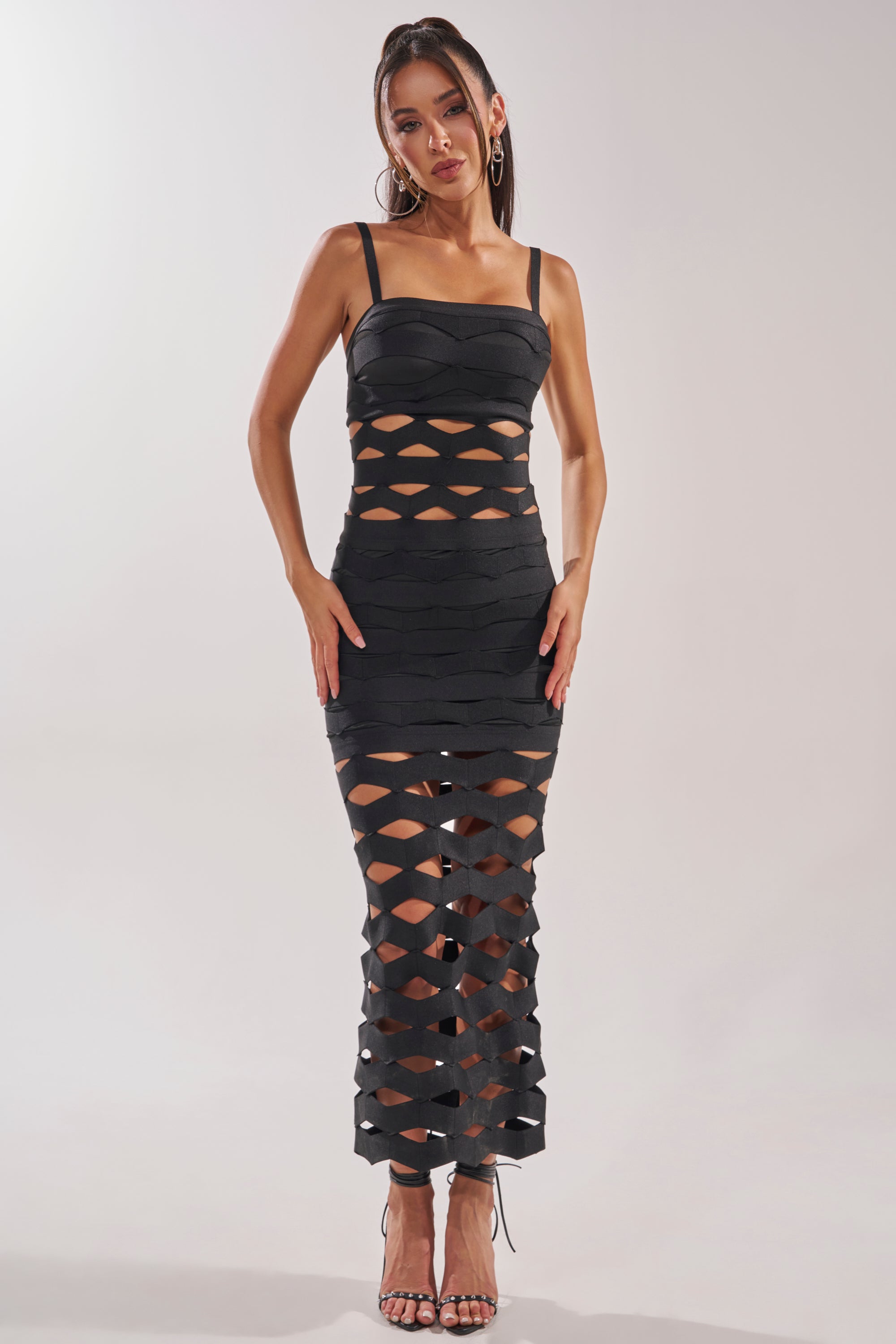 A woman stands facing forward in the STREET CHIC MAXI DRESS, featuring a black geometric cutout design, fitted top, and mermaid silhouette. The long skirt stylishly complements her strappy high-heeled sandals. 1