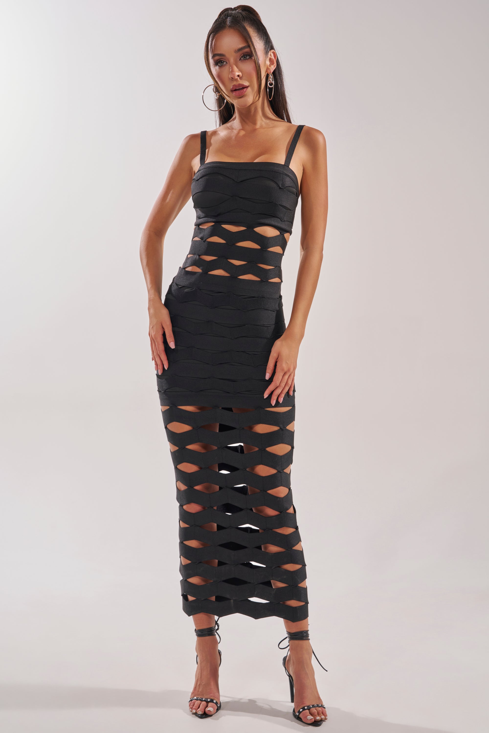 A woman poses elegantly against a plain background wearing the STREET CHIC MAXI DRESS, a black mesh maxi featuring geometric cutouts. She completes the look with long hair, hoop earrings, and high-heeled sandals. 2