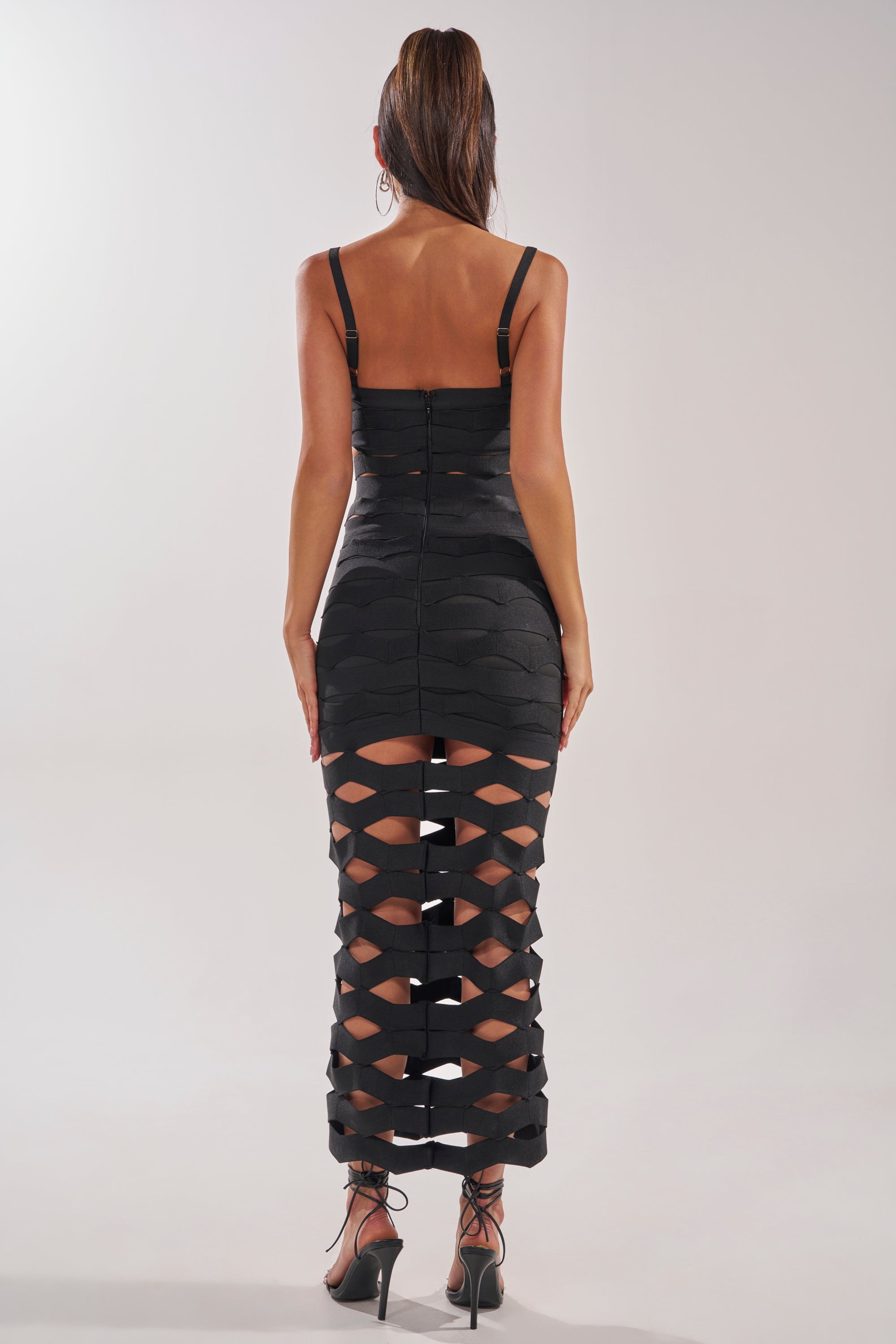 A woman faces away, modeling the STREET CHIC MAXI DRESS—a black mesh, sleeveless maxi with geometric cutouts on the lower half, a mermaid silhouette, and styled with high heels and a sleek high ponytail. 4