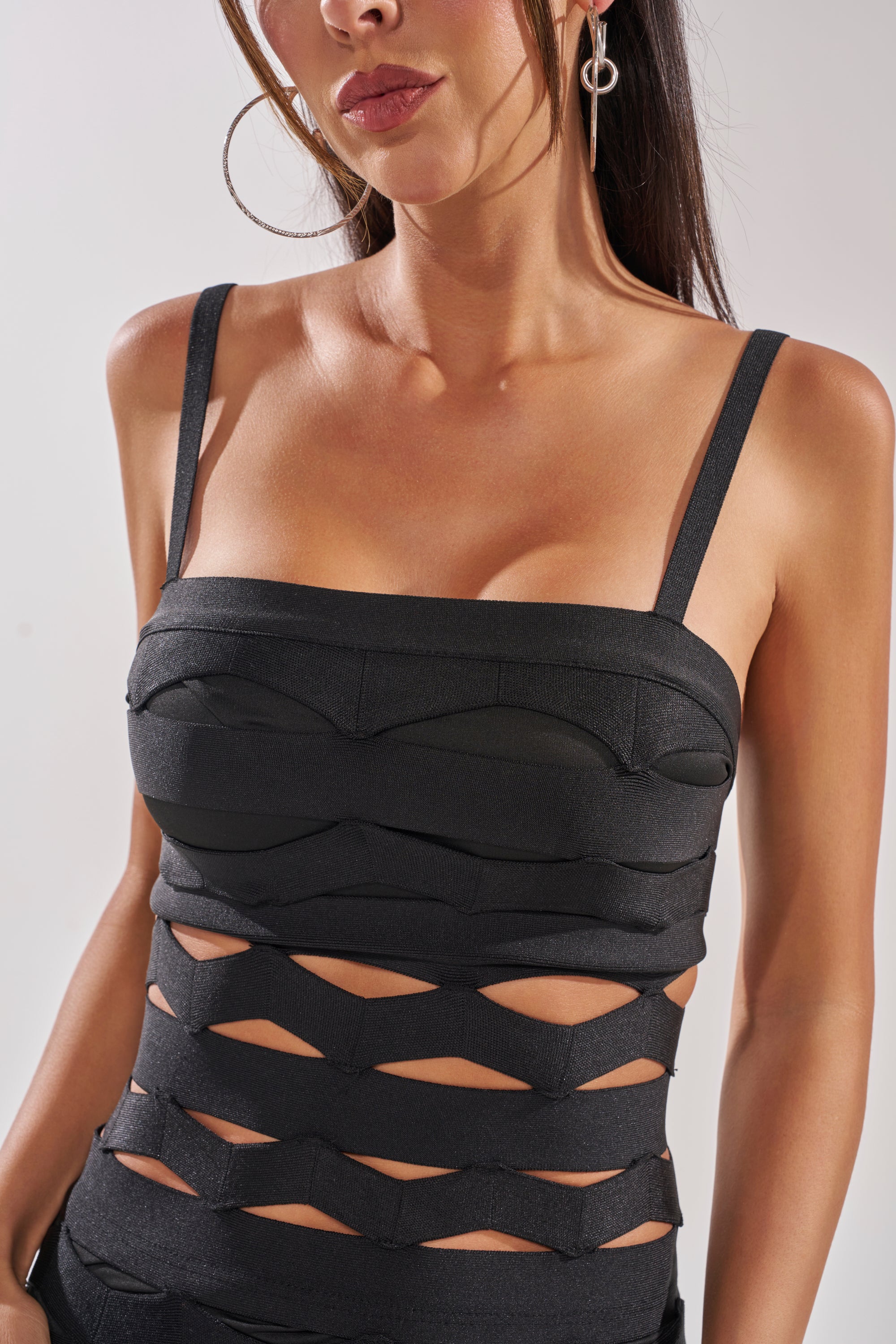 A woman with long dark hair, shown from shoulders to waist, wears the STREET CHIC MAXI DRESS—a sleeveless black geometric cutout dress with striking front details—paired with large hoop earrings. 5