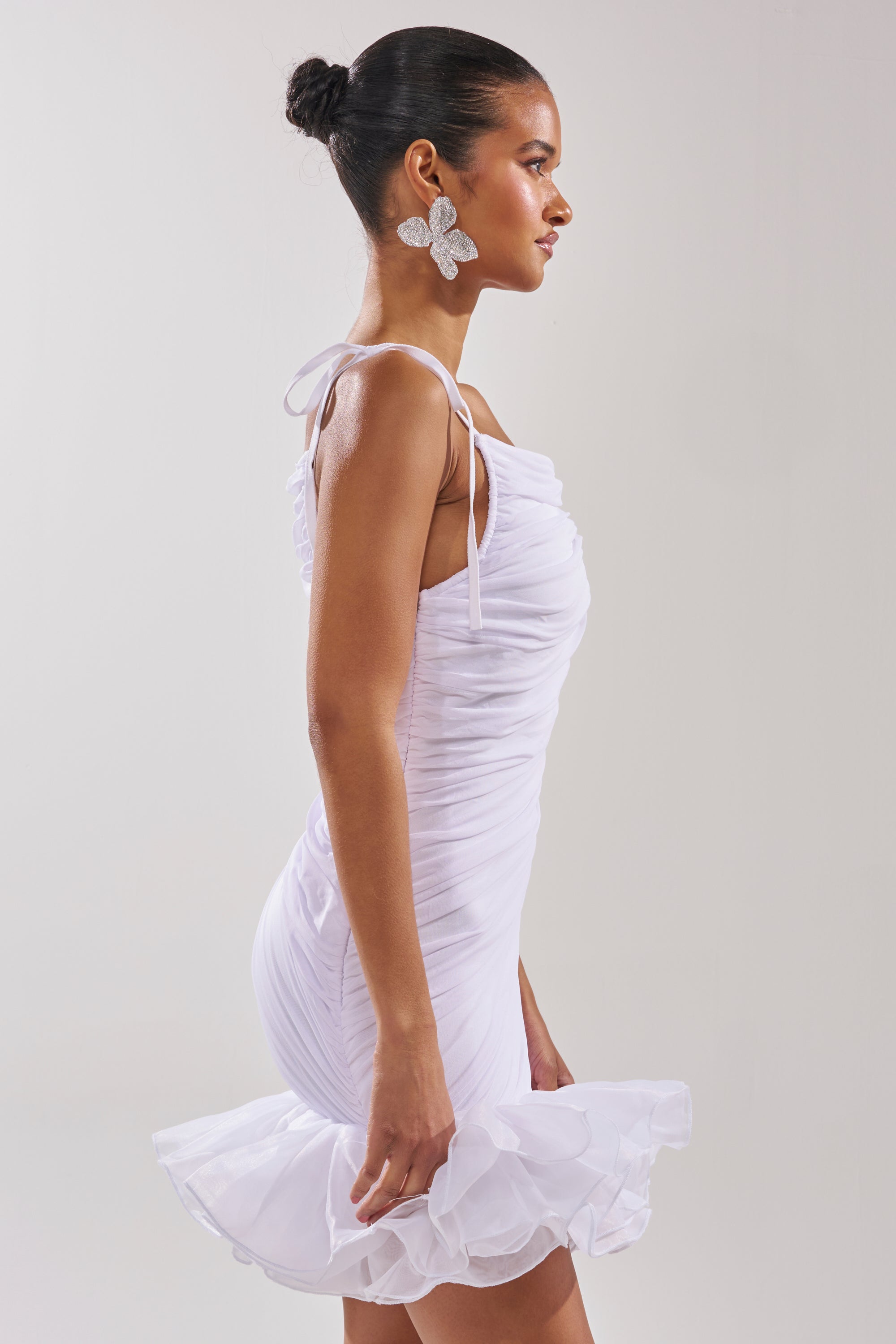 A woman with her hair in a bun models the PRINCESS PEPLUM MINI DRESS IN WHITE, featuring mesh fabric and an organza ruffle hemline, paired with butterfly-shaped earrings against a plain background. 2